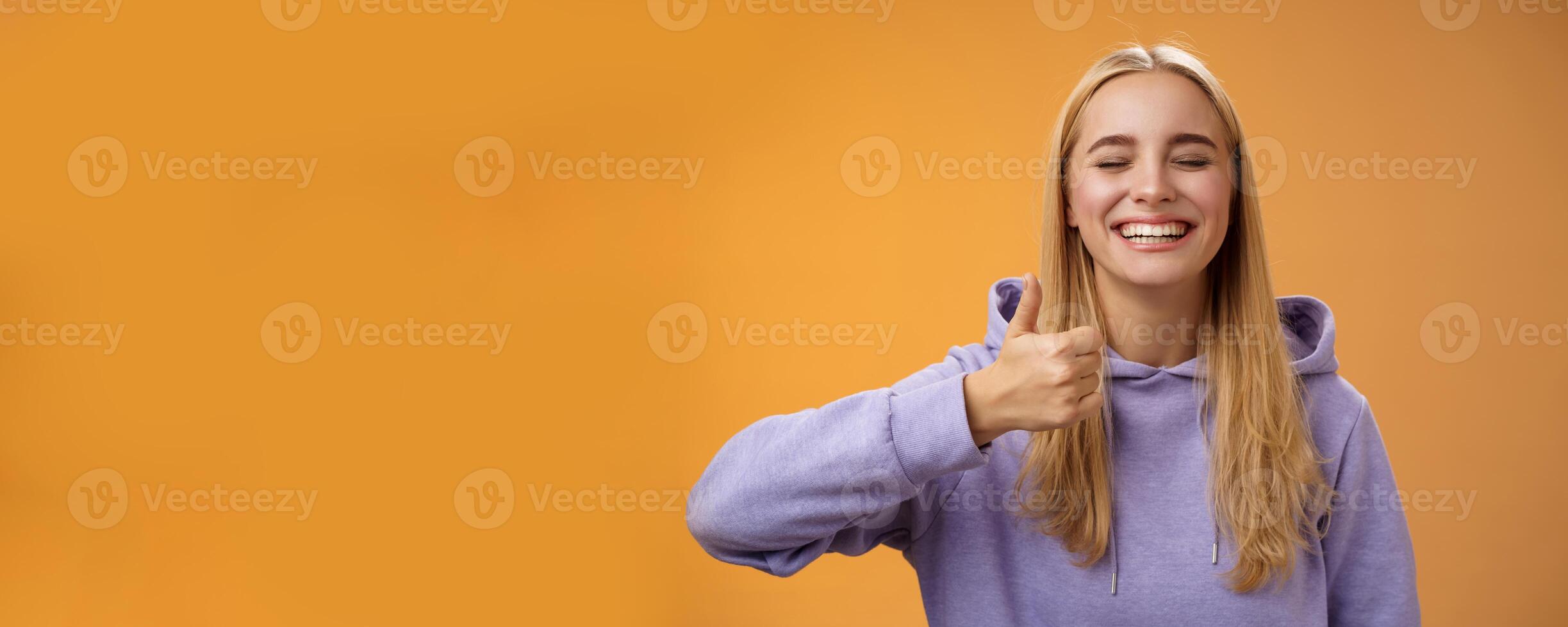 Amused joyful optimistic young excited blond european female model in hoodie smiling broadly close eyes having fun show approval gesture thumbs-up like awesome trip vacation idea, orange background photo