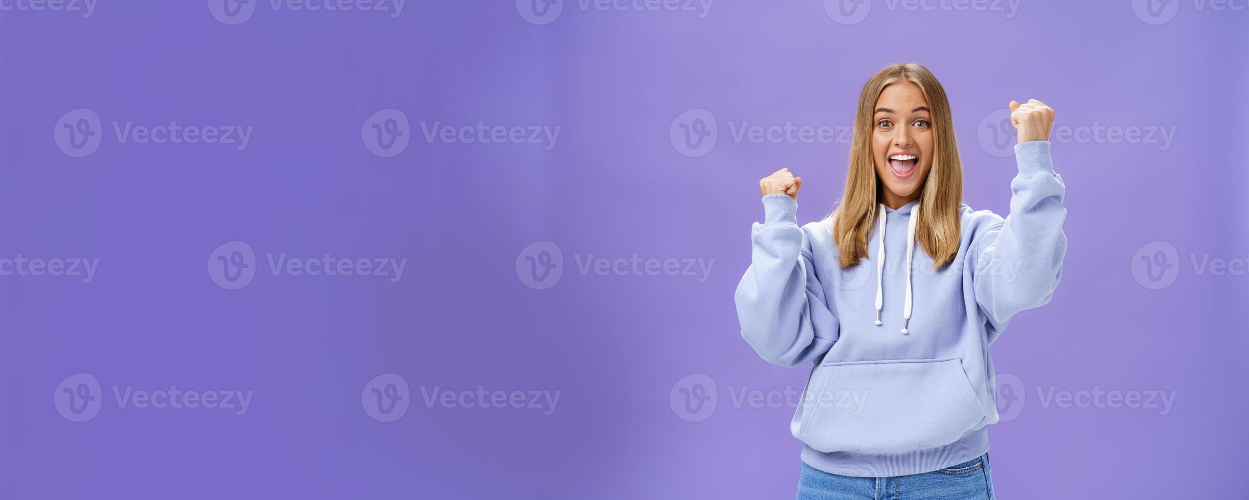 Cheerful happy and supportive young girlfriend with fair hair and tan in warm hoodide raising fists in cheer and triumph smiling saying yeah celebrating goal or success over purple background photo