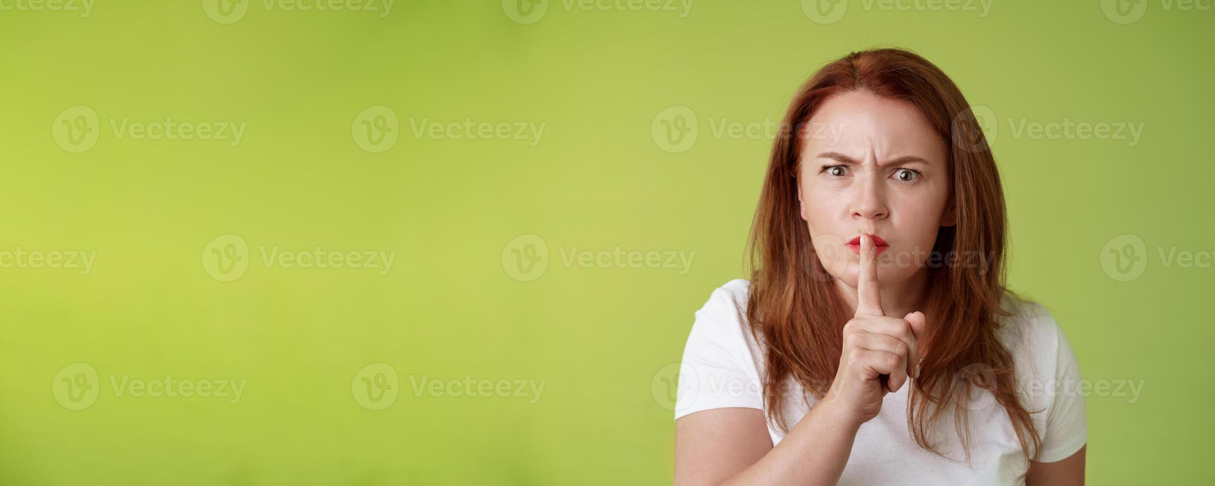 Not speak during exam. Strict serious-looking displeased middle-aged redhead woman frowning disappointed hushing say shush index finger pressed lips keep quiet gesture green background photo