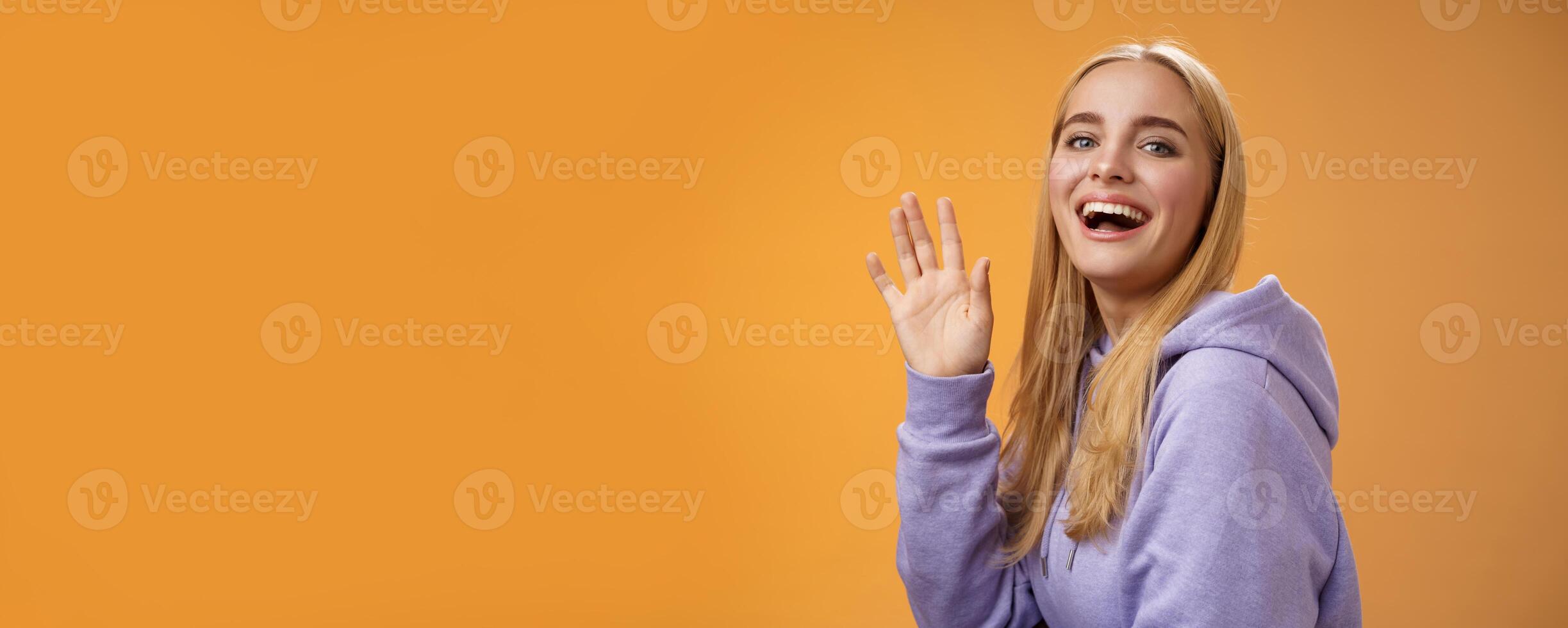 Waist-up friendly lucky cute blond girl waving palm happily smiling turning camera greet friend say hello hi see familiar person welcoming newcomers joyfully, standing orange background photo