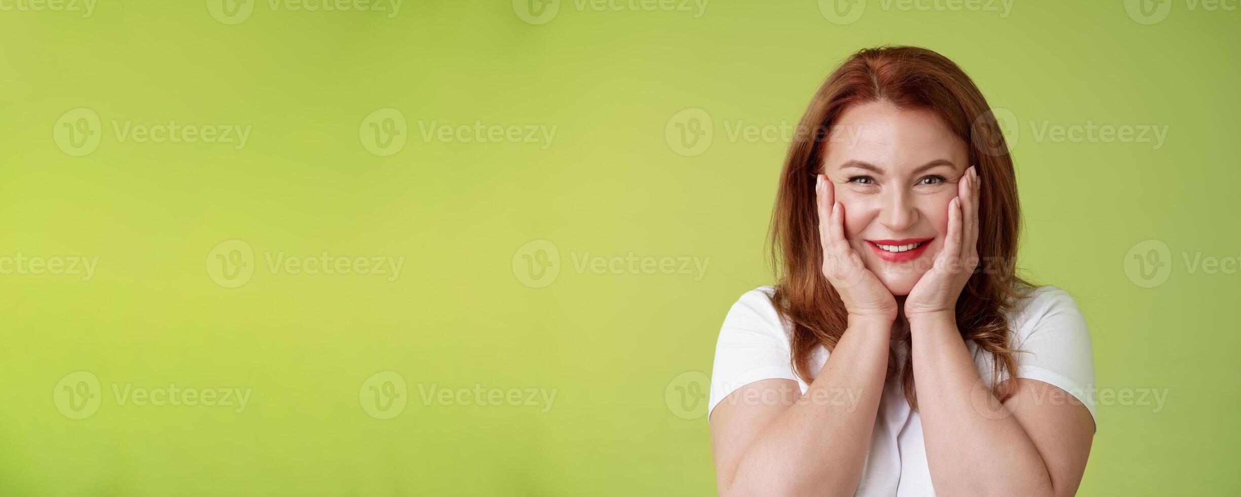 Happy delighted cheerful lucky redhead middle-aged caucasian woman blushing joyfully receive touching cute gift touch cheeks pleased delighted smiling broadly feel happiness joy green background photo