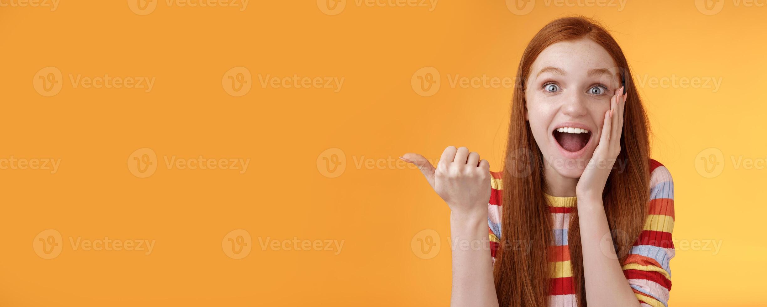 Cheerful excited impressed talkative girl pointing astonished thumb left drop jaw smiling broadly touch cheek thrilled, interesting event discuss awesome wonderful performance, orange background photo