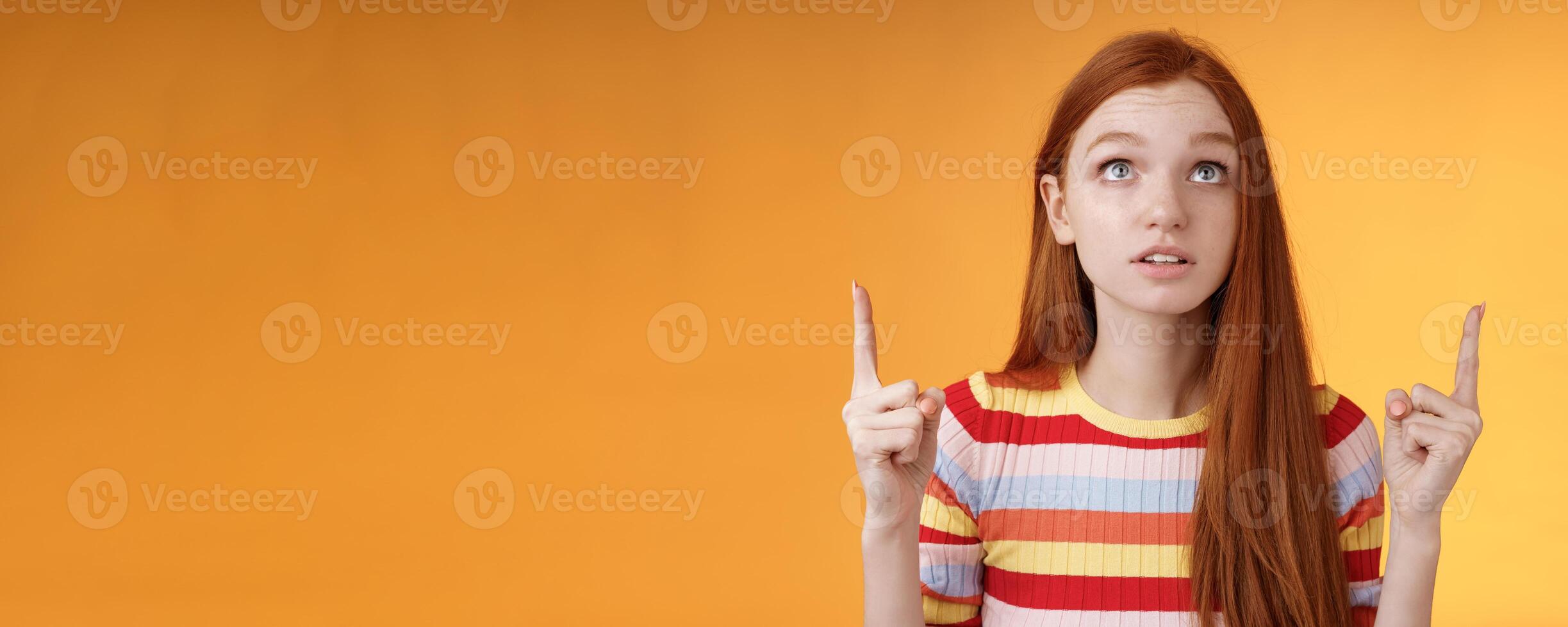 Stunned thrilled young redhead woman peer focused pointing up index fingers upwards look concentrated excited hold breath amused performance standing orange background intrigued and curious photo