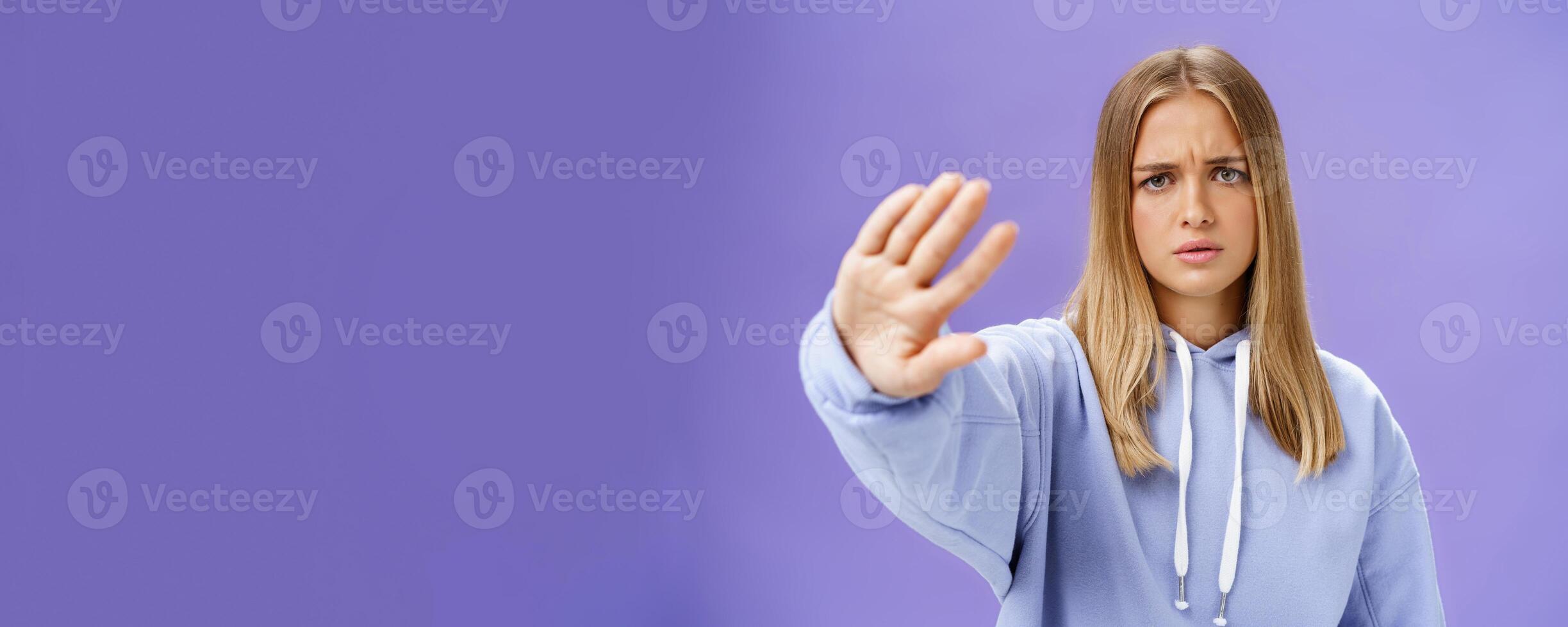 Stop photographing me. Concerned displeased young woman demanding end shooting pulling hand towards camera in no and rejection gesture frowning looking serious and displeased over blue wall photo