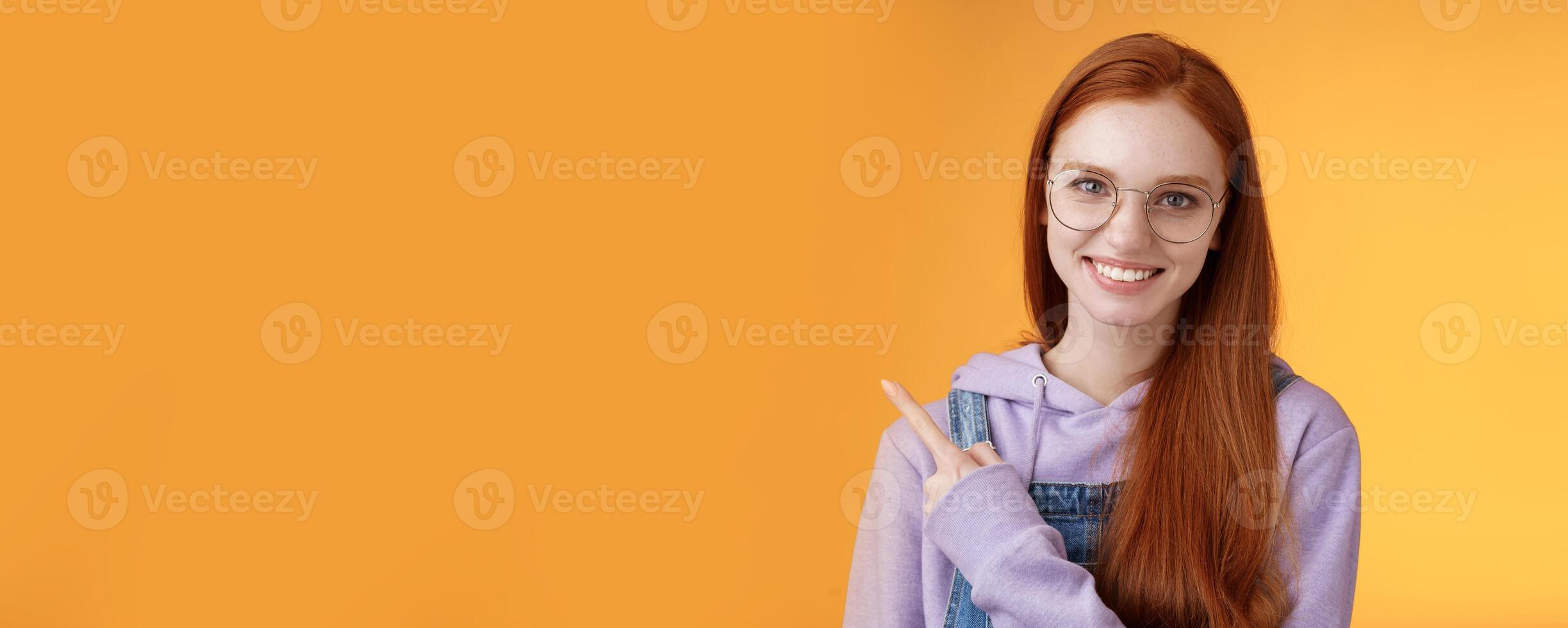 Friendly good-looking modern redhead young girl pointing left index finger showing awesome place suggesting go hang out smiling joyfully casually talking discuss new product, orange background photo