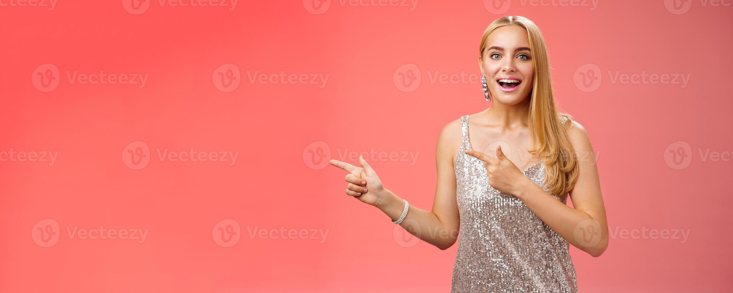 Excited carefree happy young attractive woman joyful attend awesome party in silver beautiful glittering dress pointing left suggesting come together inviting dance smiling cute red background photo
