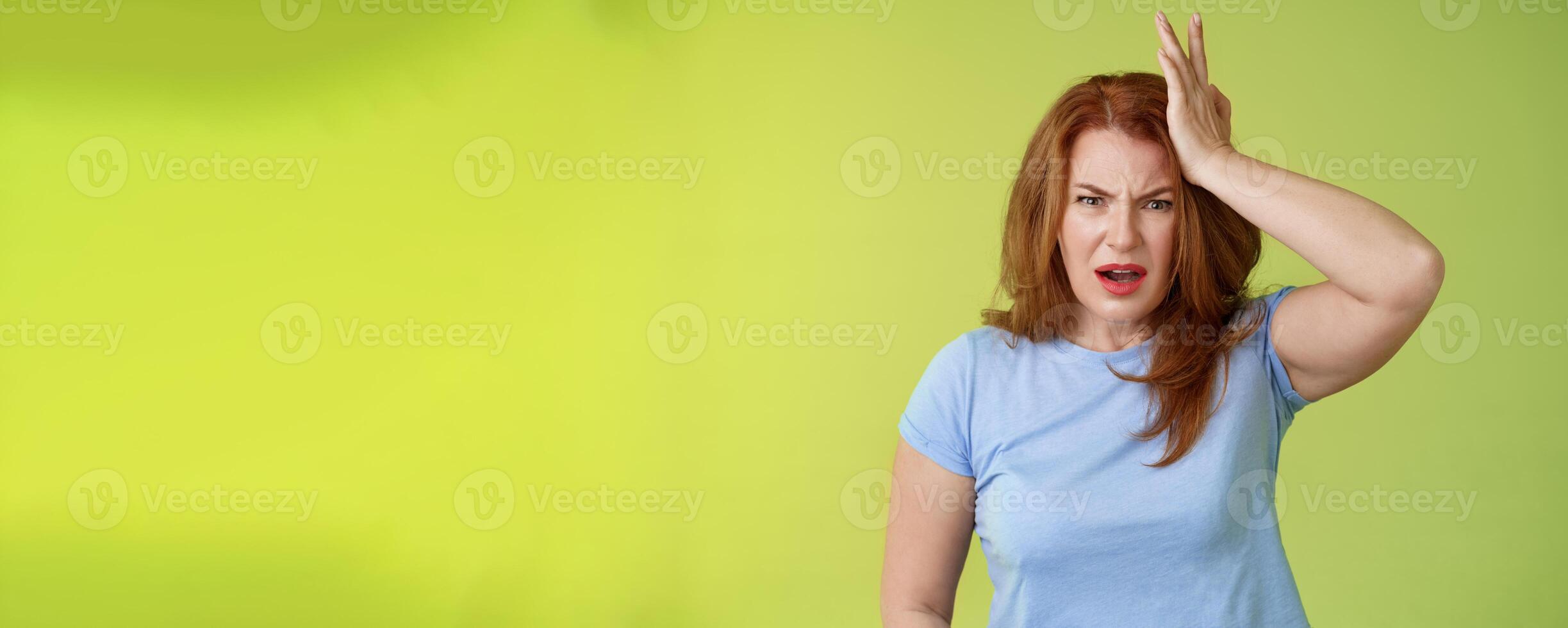 Ouch you kick my head. Frustrated concerned upset middle-aged redhead woman look shocked distressed touch temple complain kid shoot ball her face stand disappointed green background photo