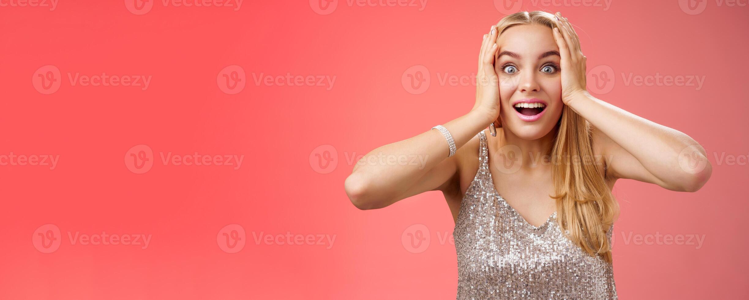Amused surprised excited young blond attractive woman in silver glittering stylish dress hold head hands widen eyes speechless look camera unbelievable thrill fan see famous person, red background photo