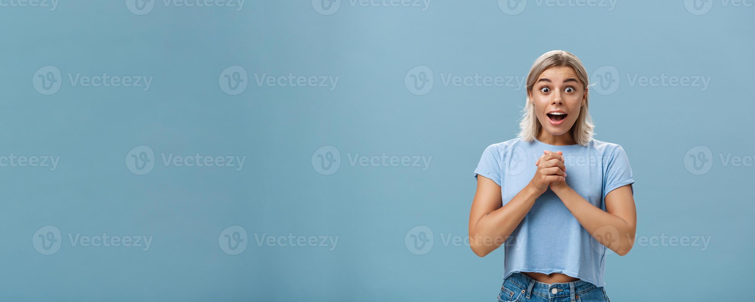 Really is it for me. Portrait of surprised grateful attractive woman with blond haircut clenching hands together on breast opening mouth gasping from admiration and fascination standing over blue wall photo