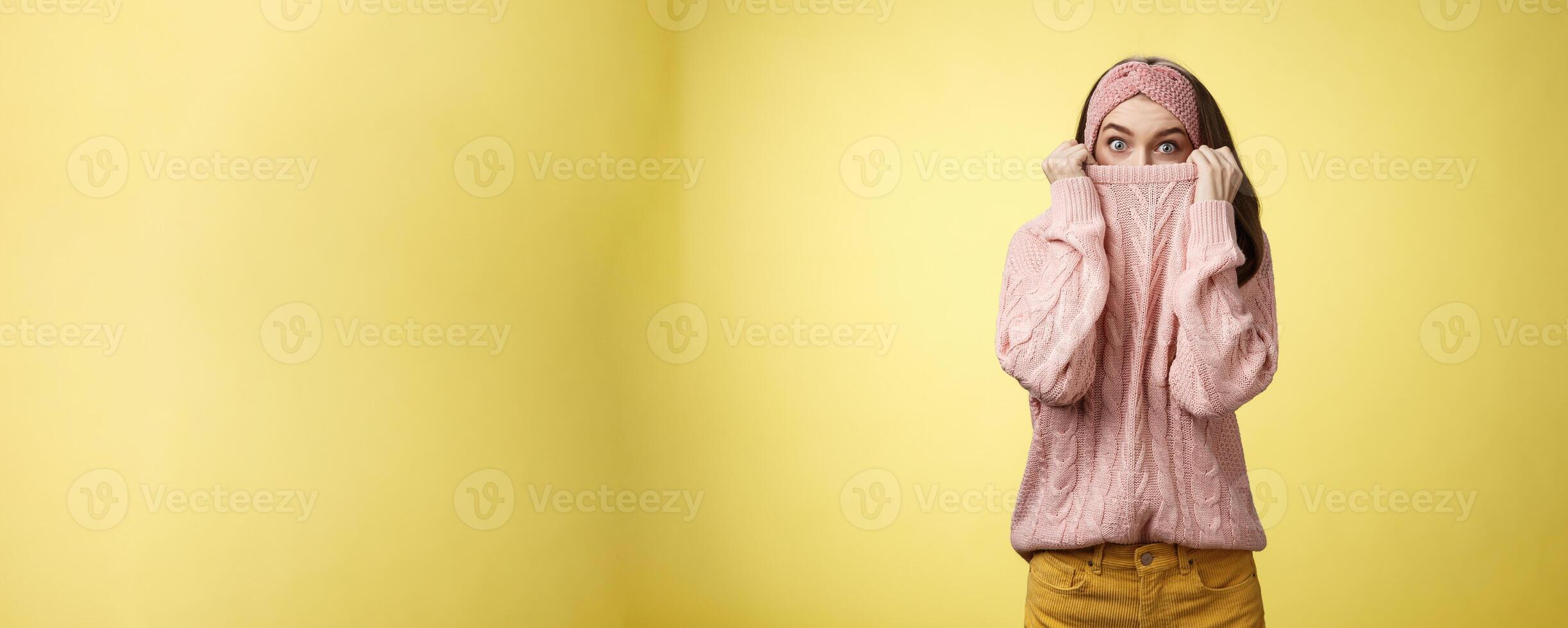 Girl hiding in collar wearing knitted sweater over nose popping eyes, staring bugged eyes at camera, hiding face reacting amazed and shocked, feeling scared, standing in stupor over yellow background photo