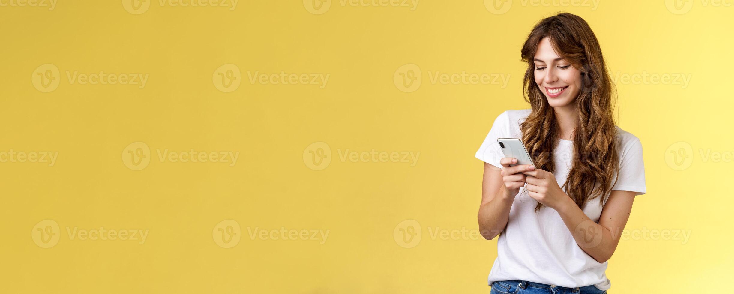 Cheerful lovely girlfriend texting friend pleased cute smile tap smartphone screen smiling broadly look mobile phone display tenderly writing post contemplate touching photo yellow background