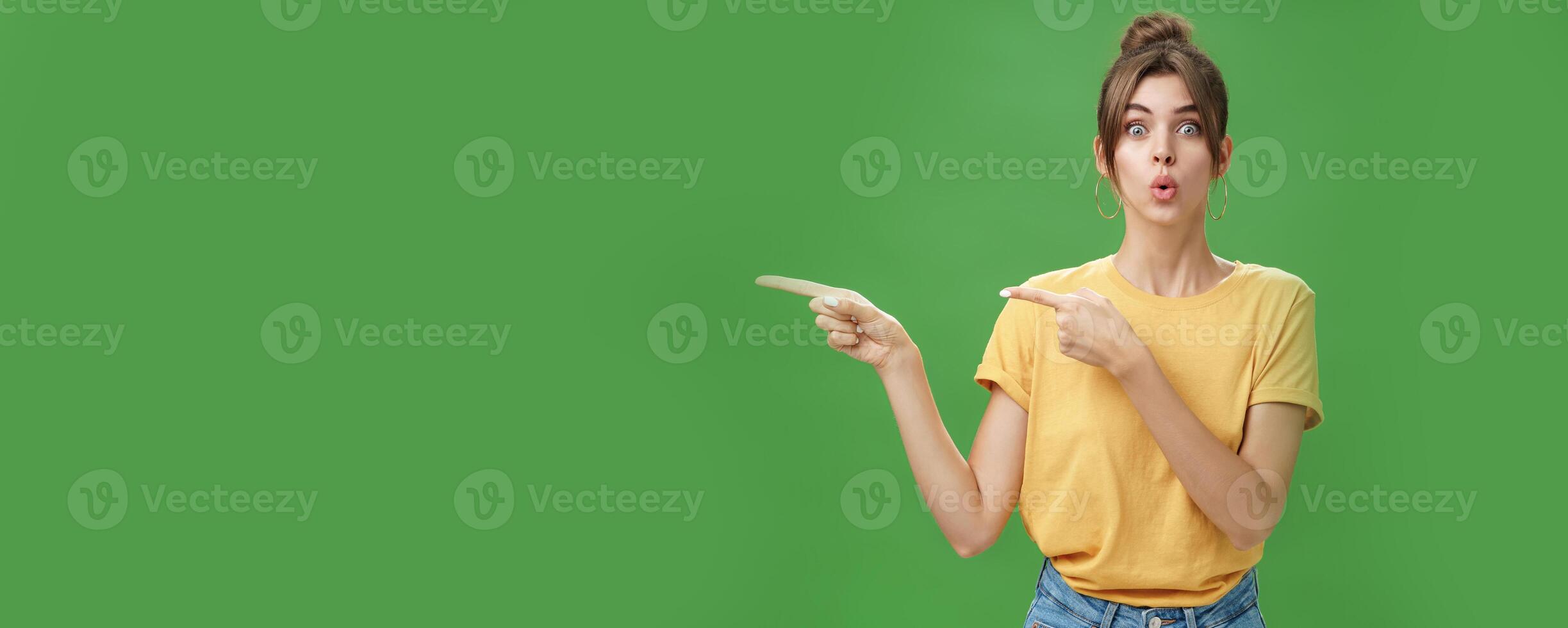 Woman reacting to awesome sales in near shop pointing left and gazing impressed and excited at camera folding lips from desire and amazement staring at camera thrilled posing over green background photo