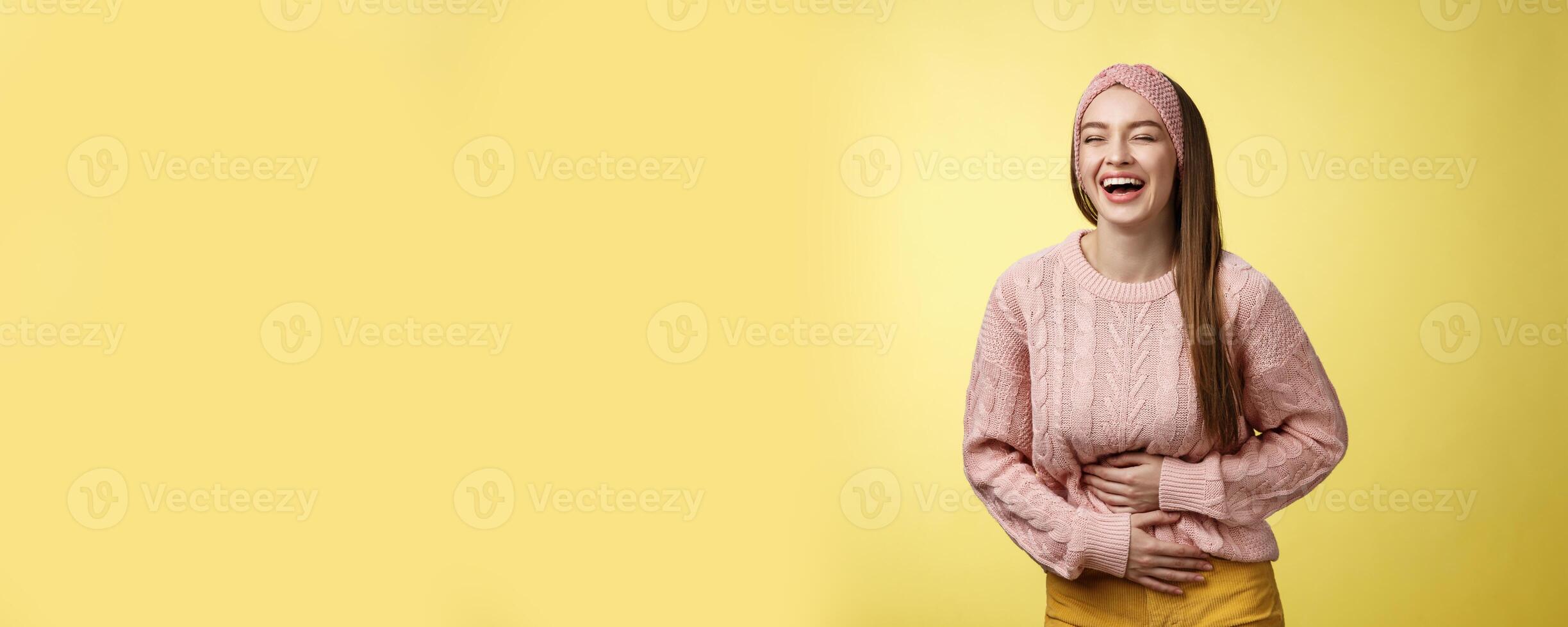 Amused and entertained charming young european female student laughing till belly hurts from giggle smiling holding palms on stomach, having fun enjoying watching funny comedy over yellow wall photo