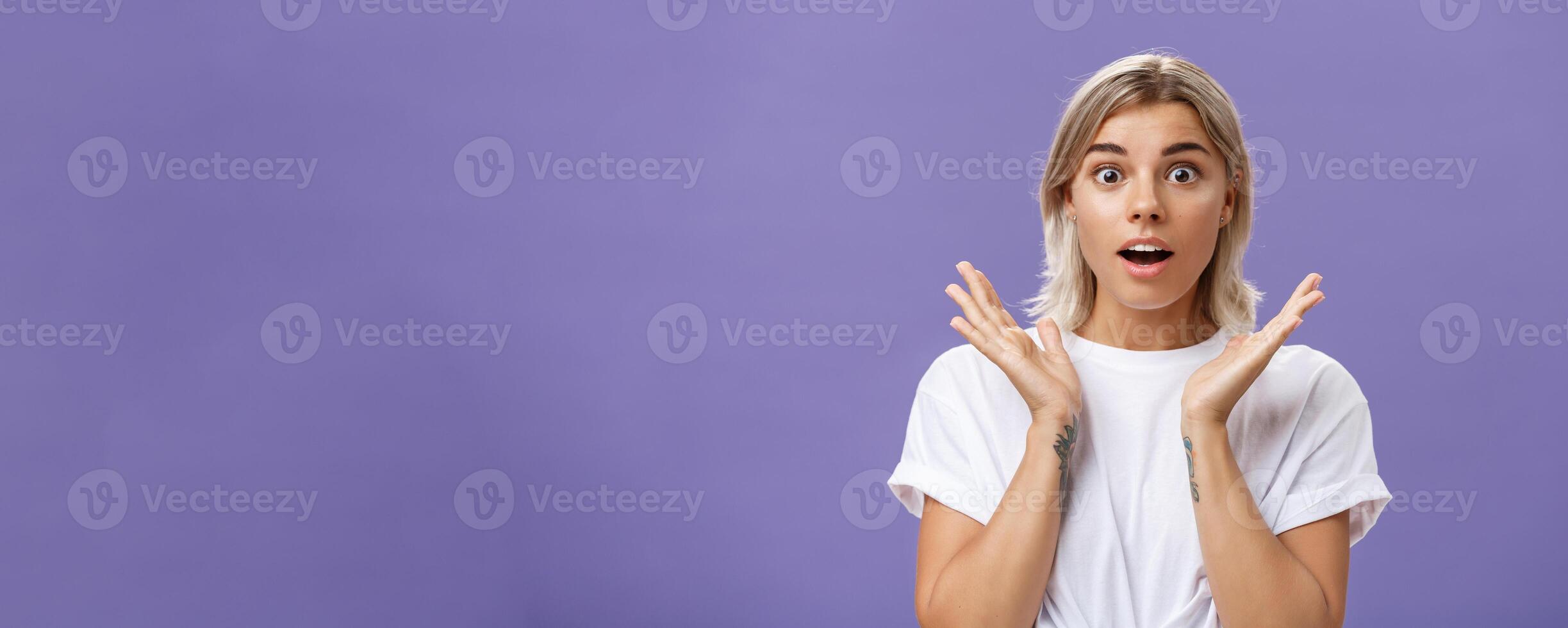 Close-up shot of charmed and impressed good-looking caucasian girlfriend with blond hair and tattoos on arms raising hands in amazed and excited gesture opening mouth and gazing mesmerized at camera photo