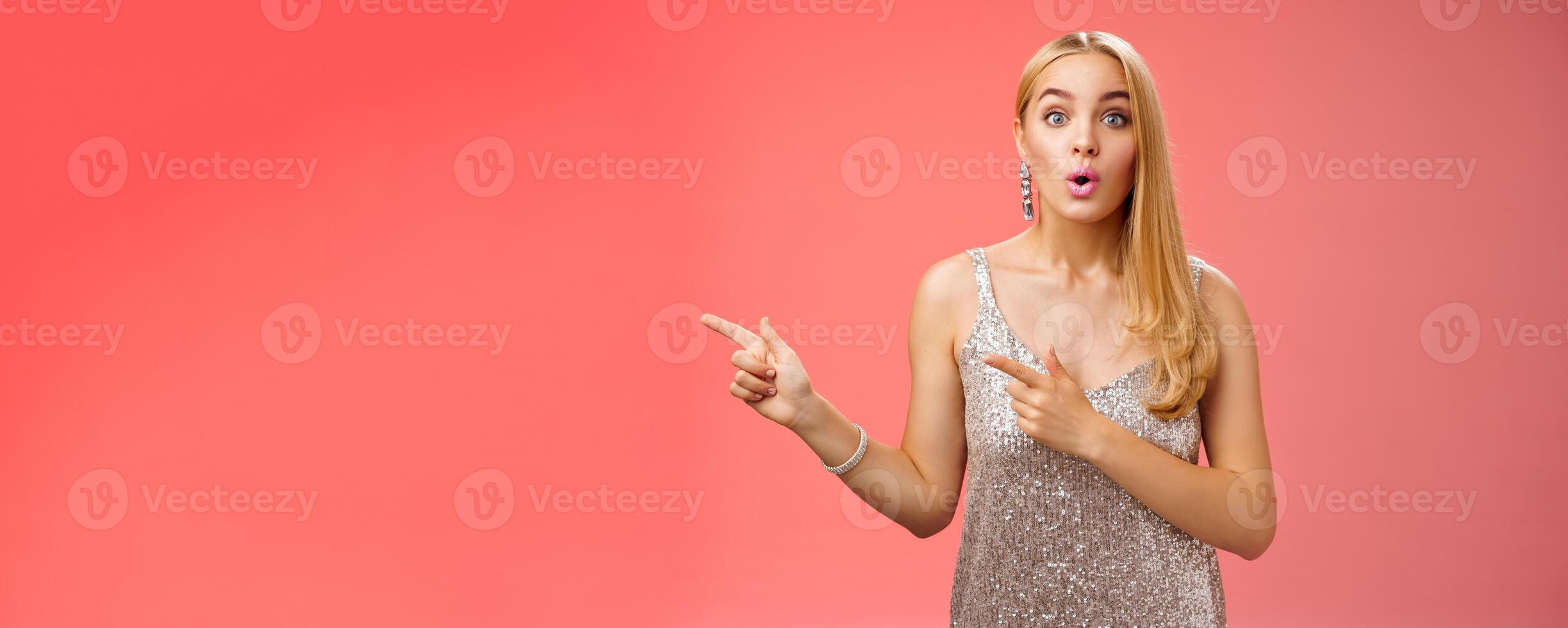 Intrigued excited attractive tender feminine blond european girl in silver stylish dress pointing left amused talking camera widen eyes thrilled asking boyfriend wanna dance shall go check out promo photo