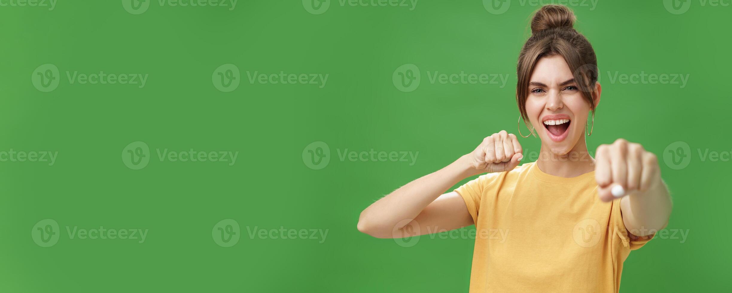 Cute female rebel in yellow t-shirt with gap teeth pulling fist towards camera as if showing fighting skills yelling daring and excited standing over green background smiling acting like boxer photo