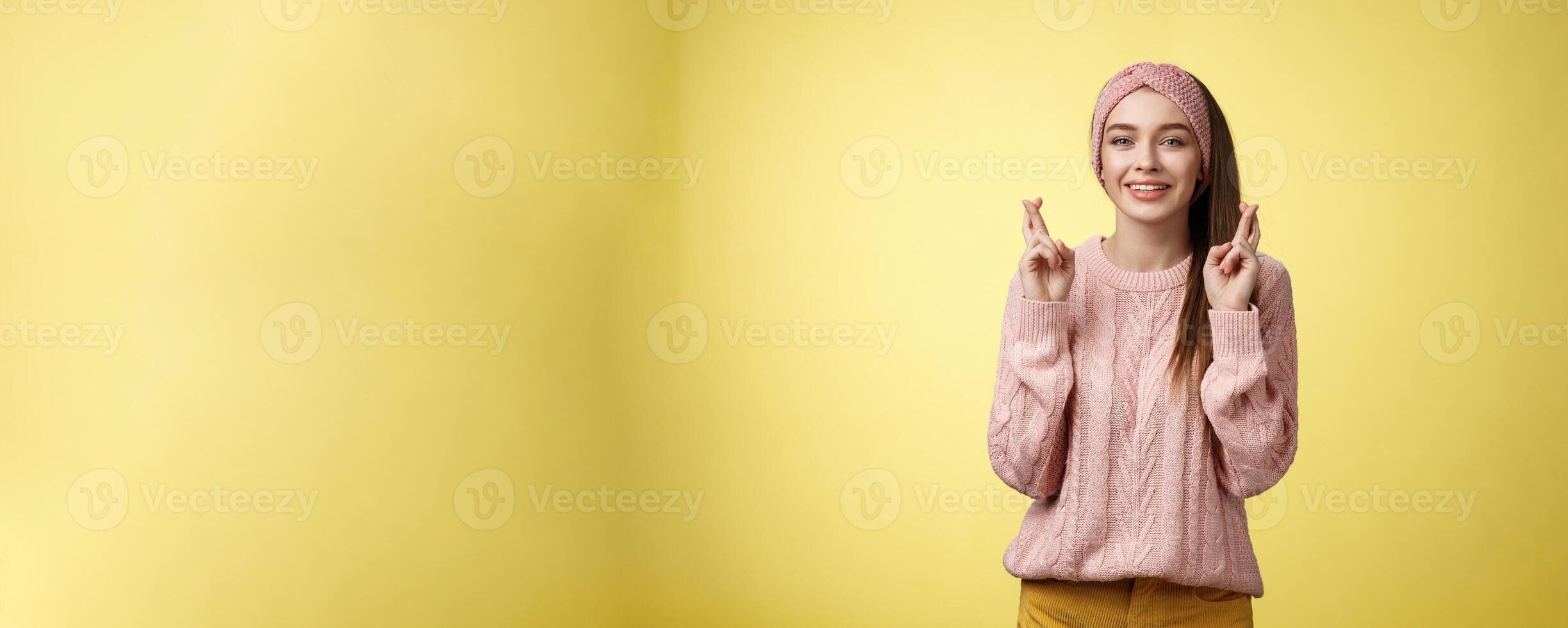 Cross my fingers for you. Supportive cute young european girlfriend wearing sweater, glamour headband making luck, wish gesture smiling hopeful having faith dream come true over yellow background photo