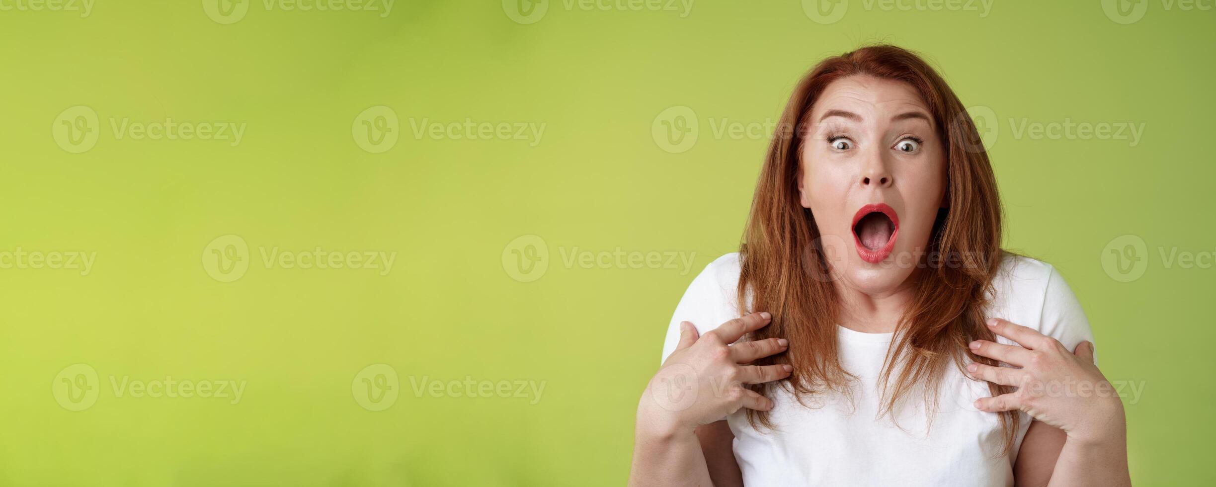 Shocked panicking redhead middle-aged woman gasping drop jaw open mouth stare camera freak-out anxious pointing herself impressed terrified frustrated nervously react green background photo