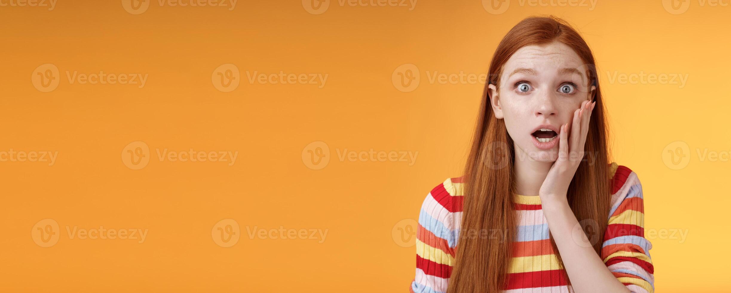 Concerned worried ginger girl blue eyes drop jaw gasping touch cheek confused looking nervously anxious, show empathy hearing terrible upsetting story standing orange background photo