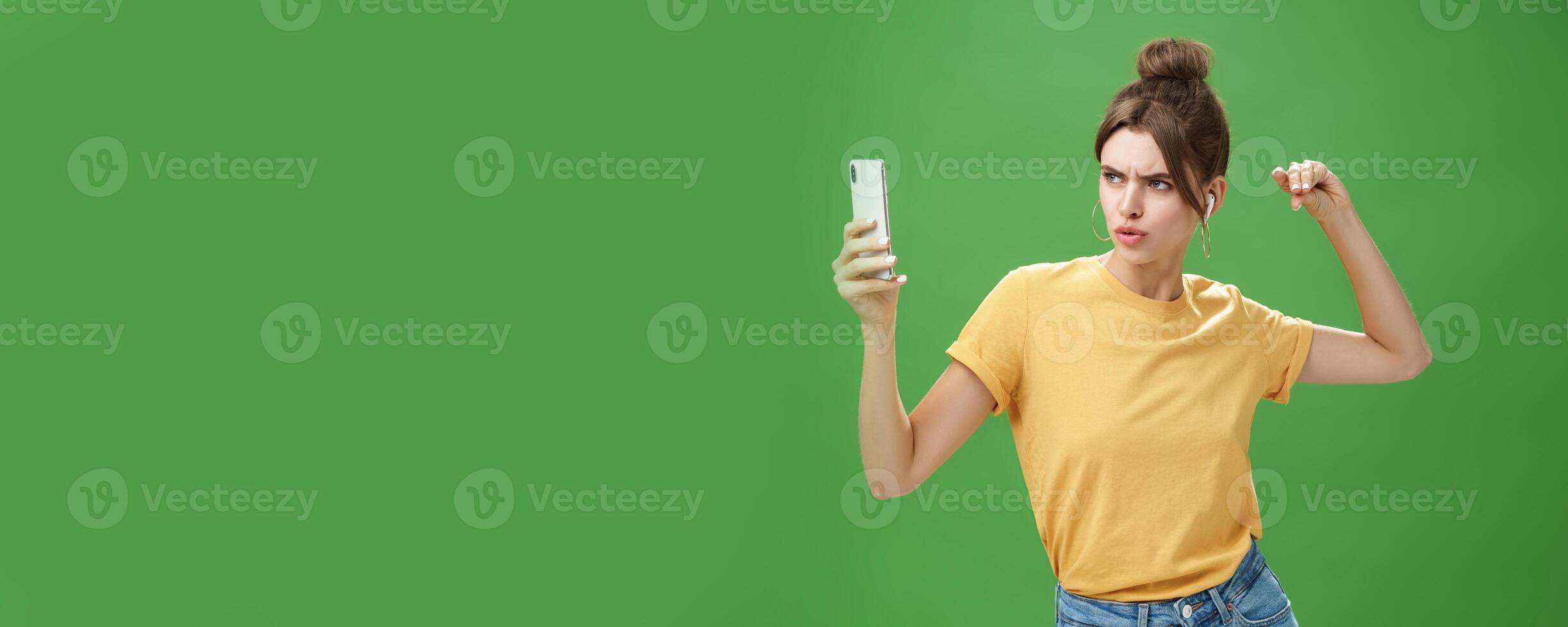 Woman making serious look to take selfie while listening music in wireless earphones posing and staring at smartphone screen daring and aggressive, liking post photos online over green background
