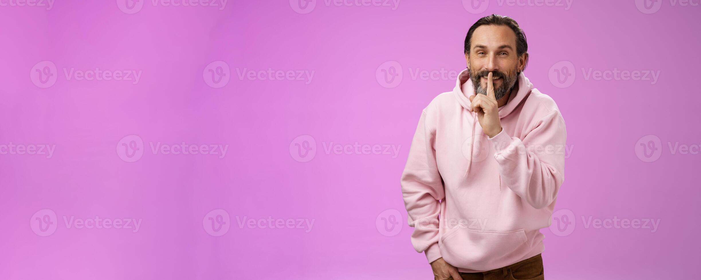 Lips sealed secret. Portrait alluring cheeky stylish adult bearded guy in pink hoodie asking keep quiet show shush shh gesture index finger pressed mouth smiling devious smug face, have idea photo