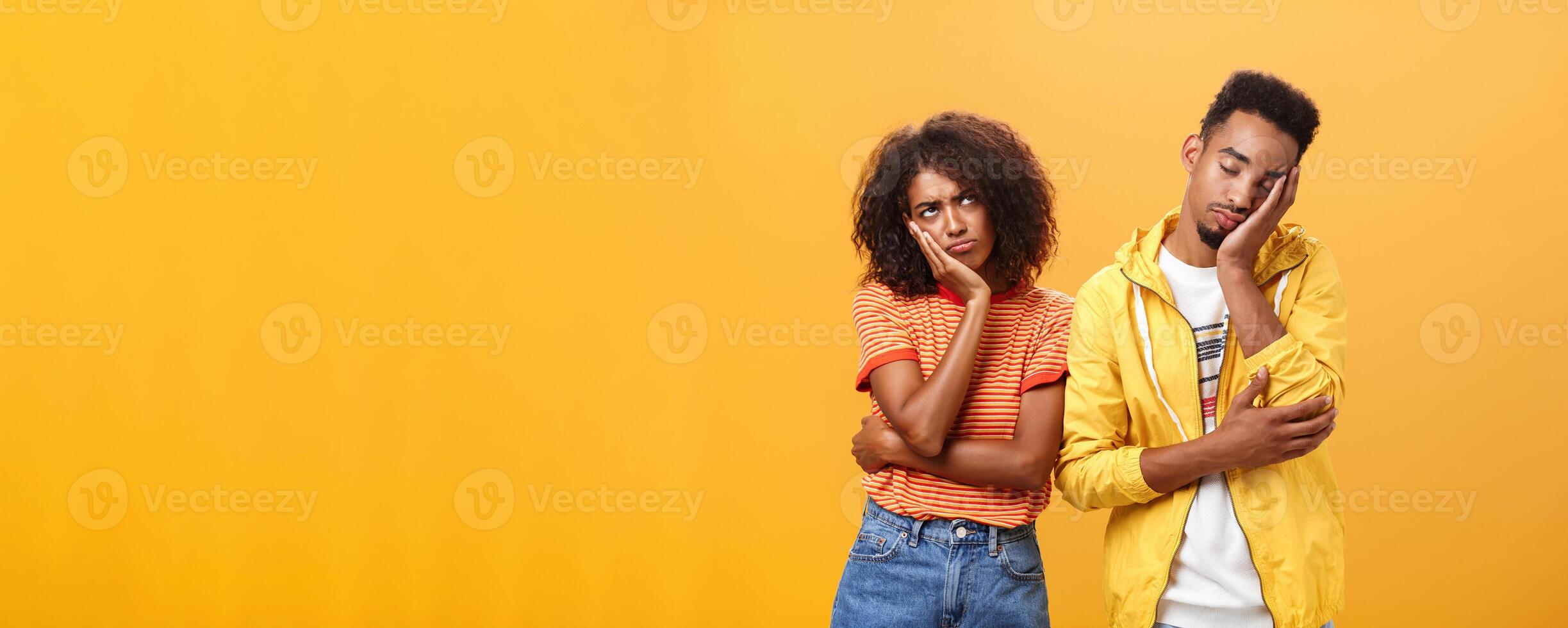 Girl feeling displeased and offended on boyfriend who fell asleep during date pursing lips frowning looking up while boyfriend leaning head on face with closed eyes and tired look over orange wall photo