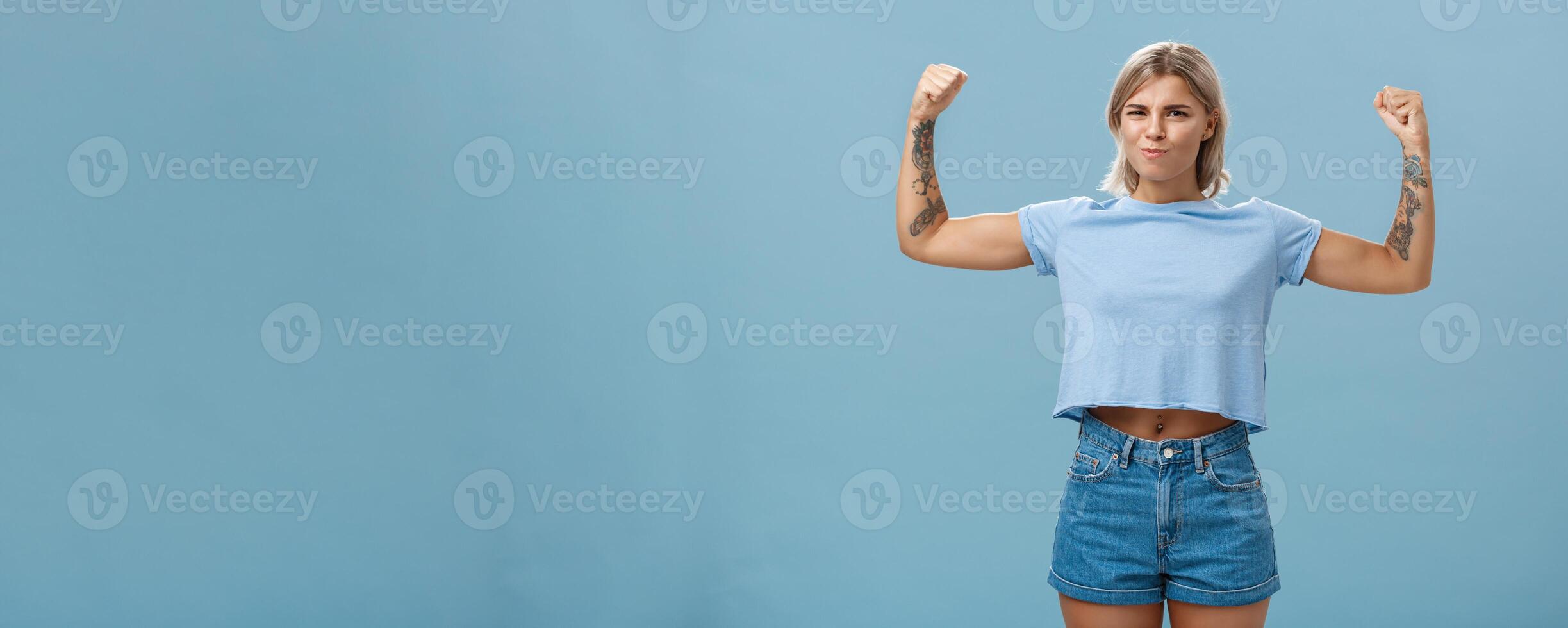 Strong and powerful good-looking sportswomen with tattoos in t-shirt and shorts raising arms showing big muscles and biceps smiling proudly while bragging with physical strength over blue wall photo