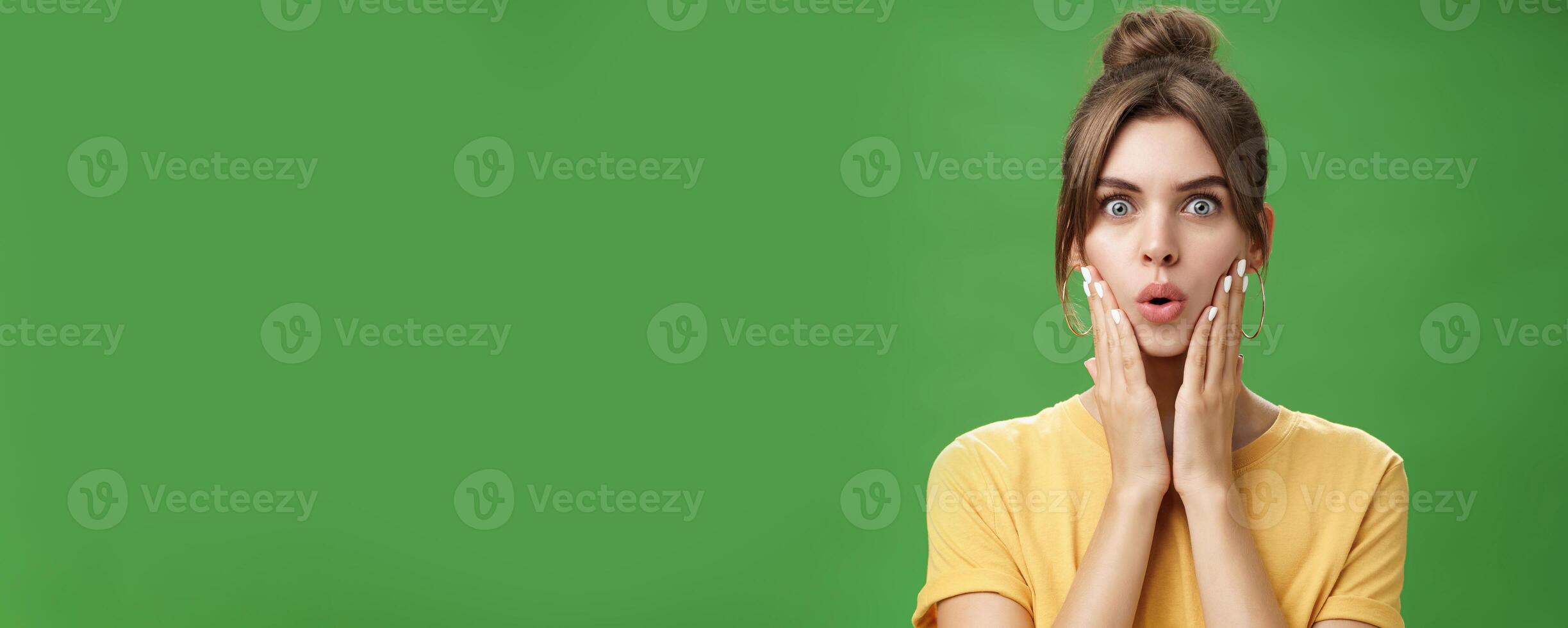 Portrait of impressed and shocked cute feminine woman in yellow t-shirt folding lips from excitement and interest touching cheeks surprised reacting to amazing rumor posing over green background photo