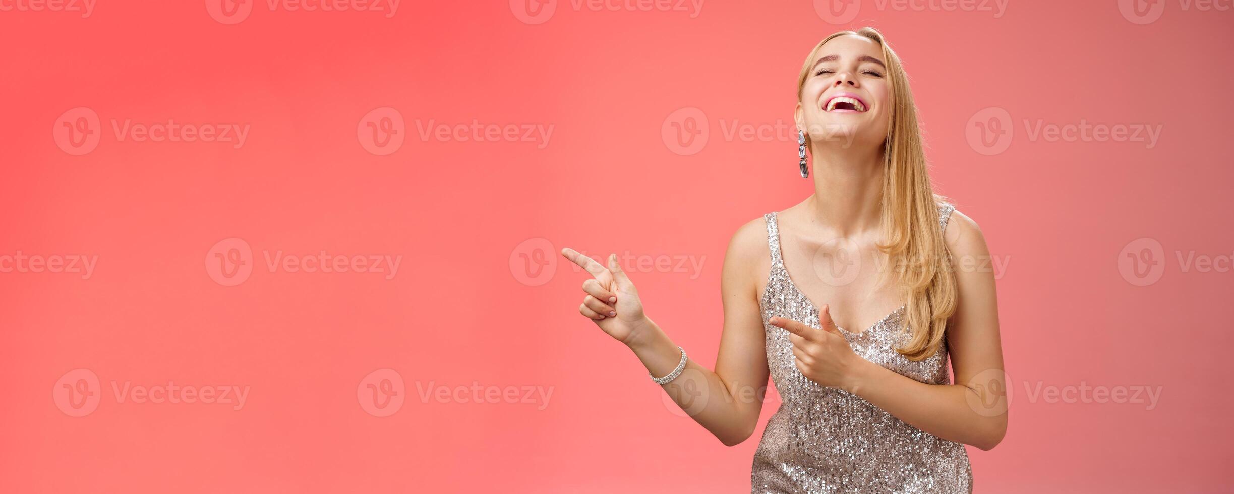 Funny carefree blond european woman in silver glittering evening dress raise head up close eyes laughing happily pointing left see hilarious person joking fool around, standing red background photo