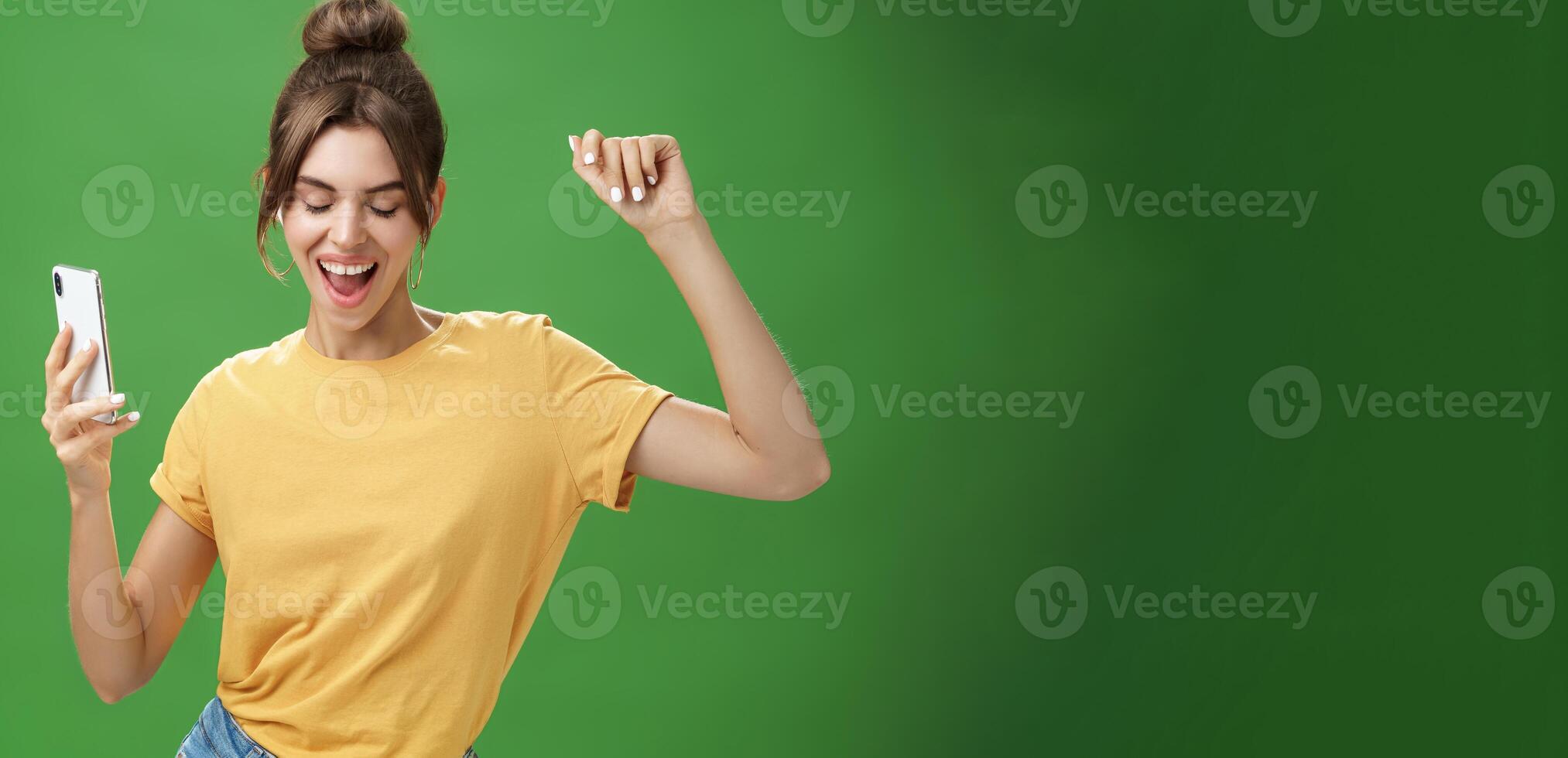 Cool and stylish happy woman with tattoo and gap teeth in yellow t-shirt dancing joyfully listening music in wireless earphones holding smartphone closing eyes and smiling from delight over green wall photo