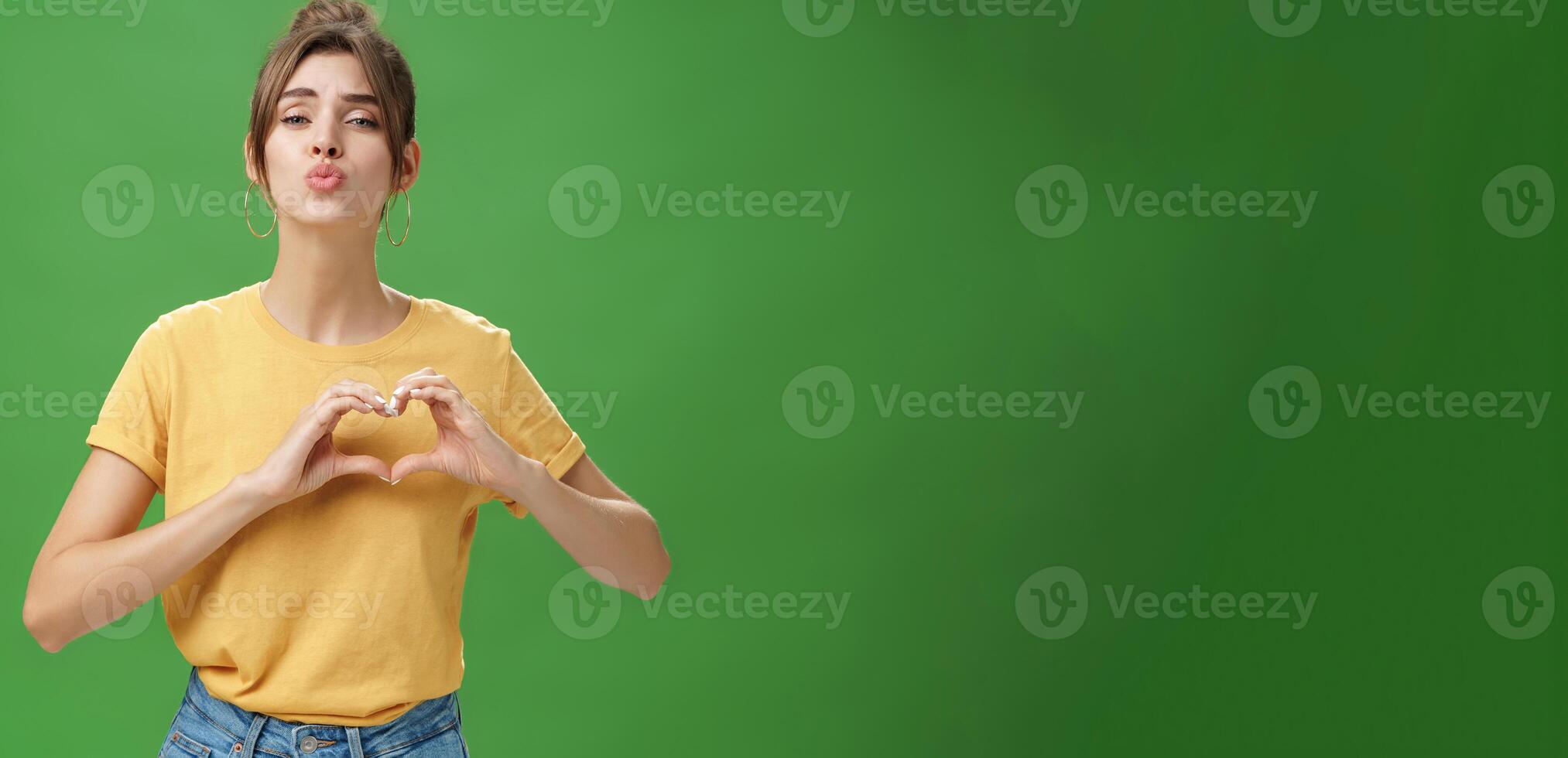 Sensual charming woman in yellow t-shirt and combed hairstyle in yellow t-shirt showing heart gesture against chest folding lips to give kiss or mwah tender and loving over green background photo