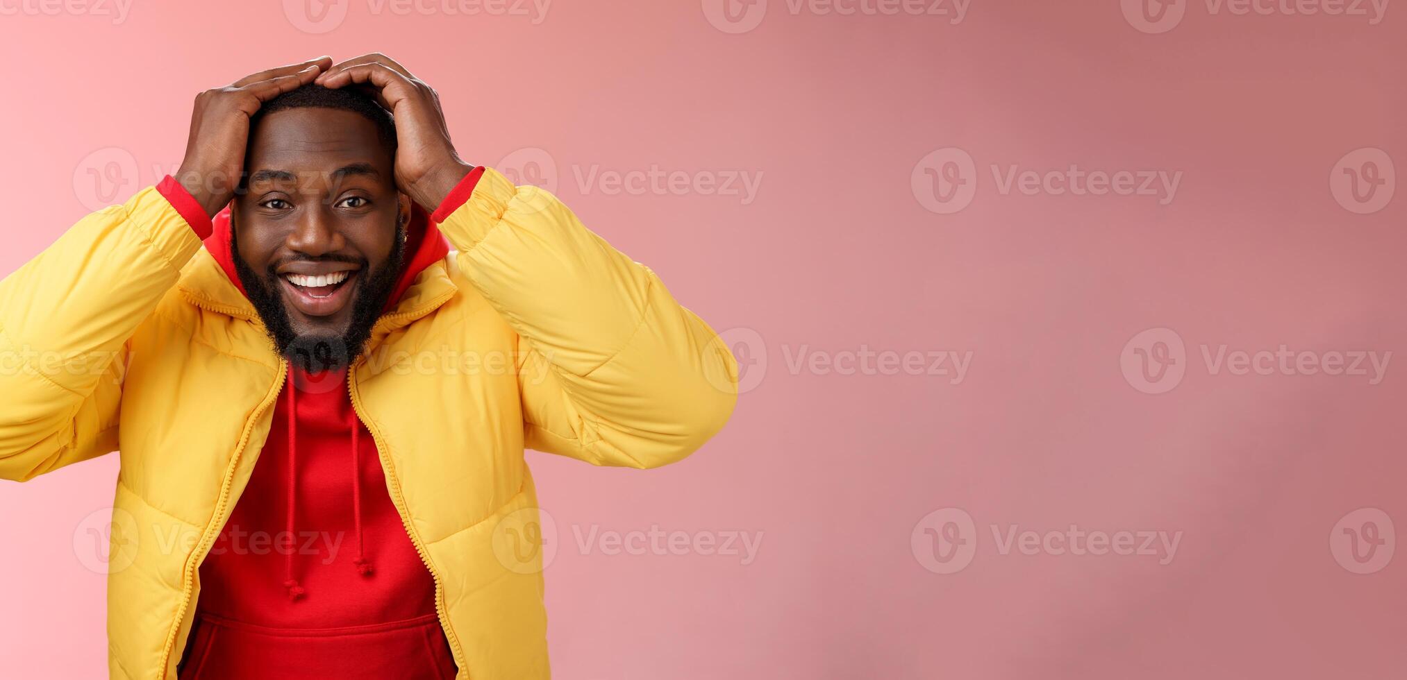 Waist-up surprised happy lucky young african-american bearded male in yellow jacket holding hands head smiling impressed joyfully receive awesome proposal dream come true, pink background photo