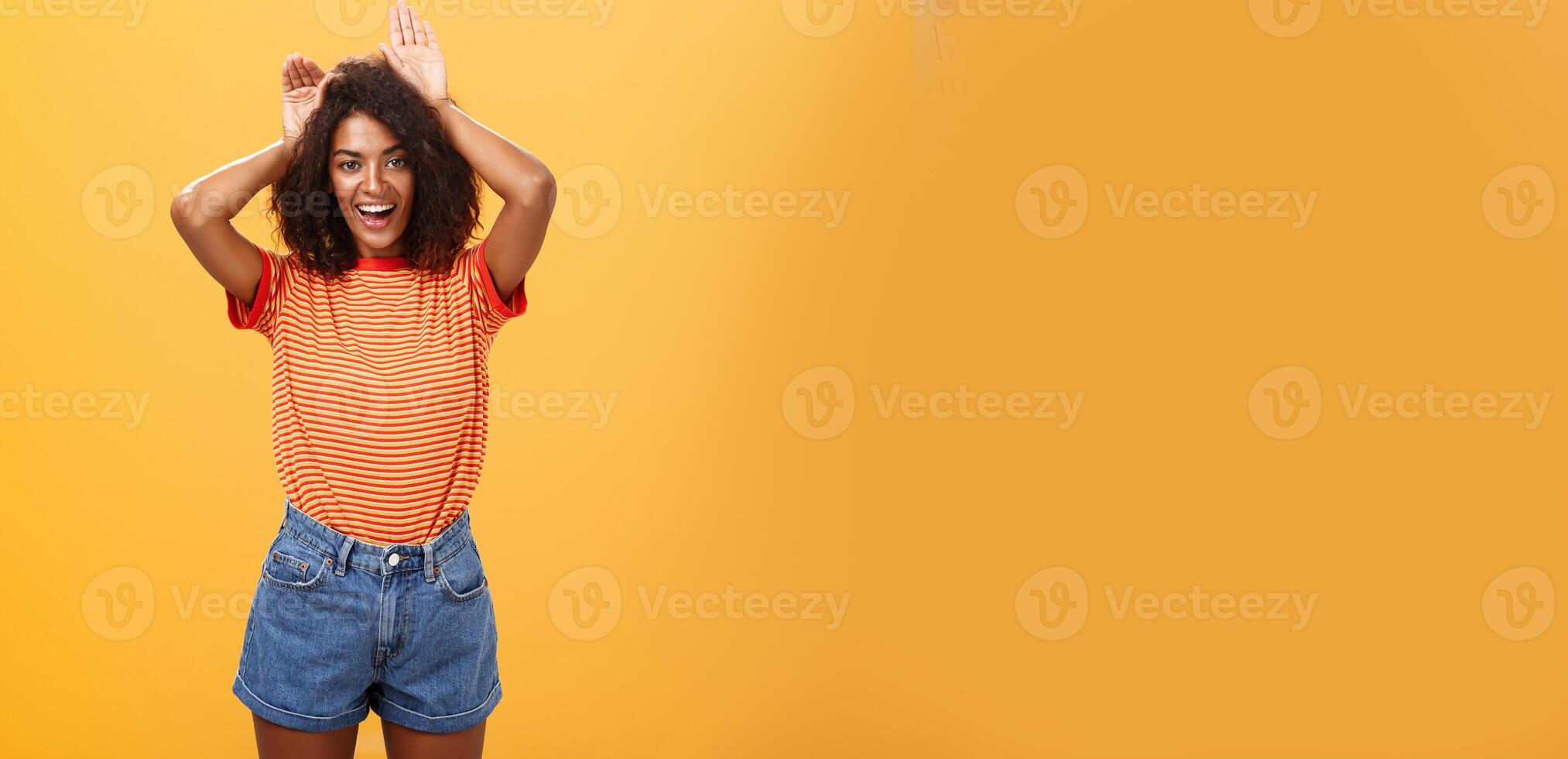 Let me be your bunny. Portrait of charming enthusiastic and charismatic happy dark-skinned female with afro hairstyle holding palms on head like animal ears making cute face over orange background photo