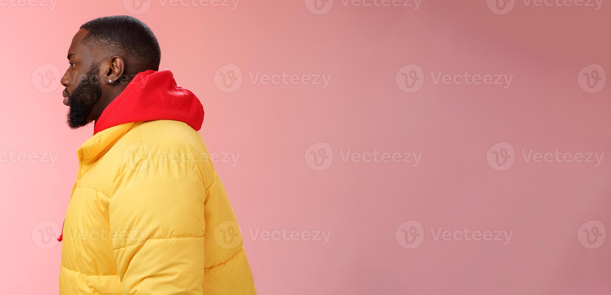 Profile studio shot bearded young 25s african guy in yellow jacket red hoodie look left normal unbothered relaxed expression standing queue order fastfood pink background, waiting take-away photo