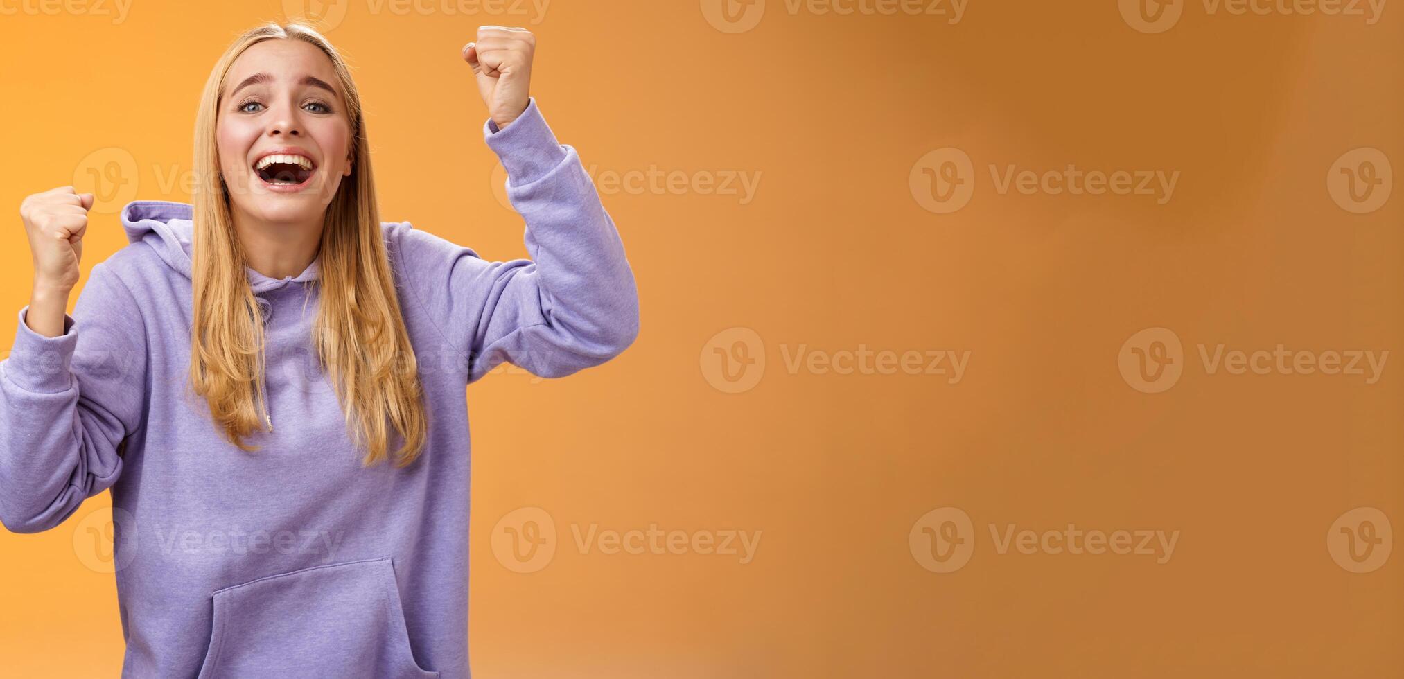 Happy cheerful young supportive woman cheering sister win smiling dedicated fun hope favorite team score goal raising clenched fists yelling happily celebrating triumphing victory, orange background photo