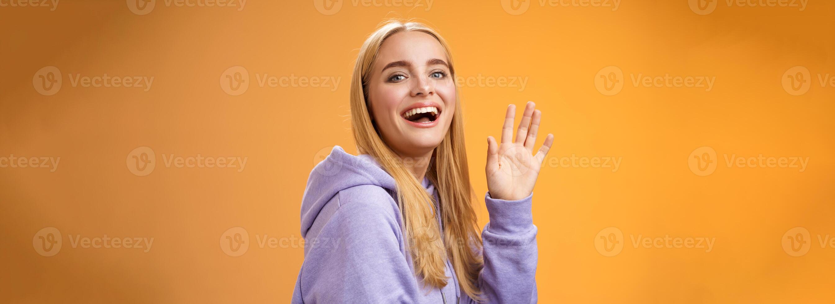 Waist-up friendly lucky cute blond girl waving palm happily smiling turning camera greet friend say hello hi see familiar person welcoming newcomers joyfully, standing orange background photo