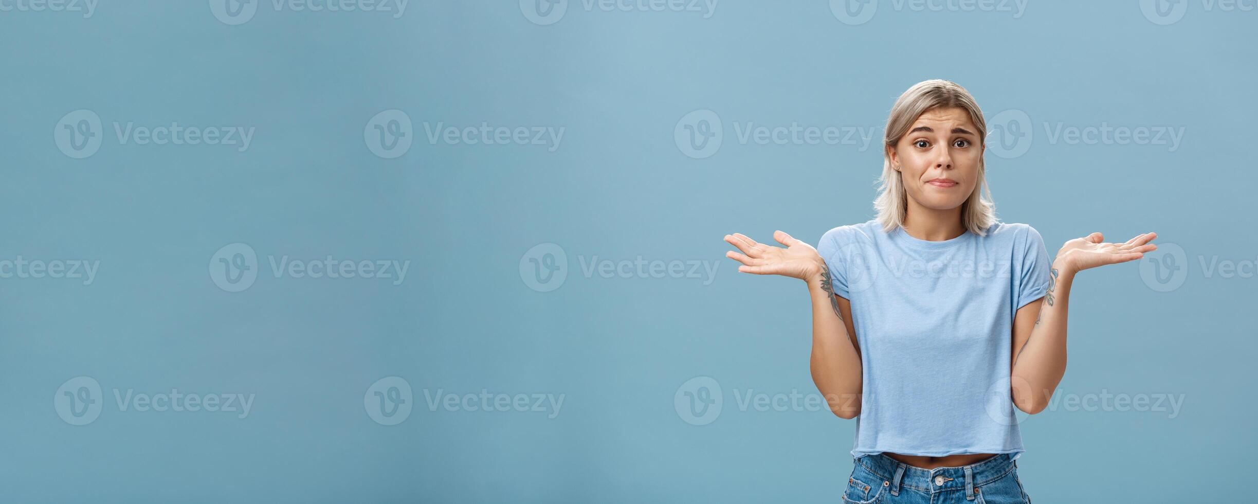 No idea how help sorry. Portrait of unsure gloomy girlfriend feeling regret shrugging with hands near shoulders frowning and pursing lips being uncertain and unaware over blue background photo