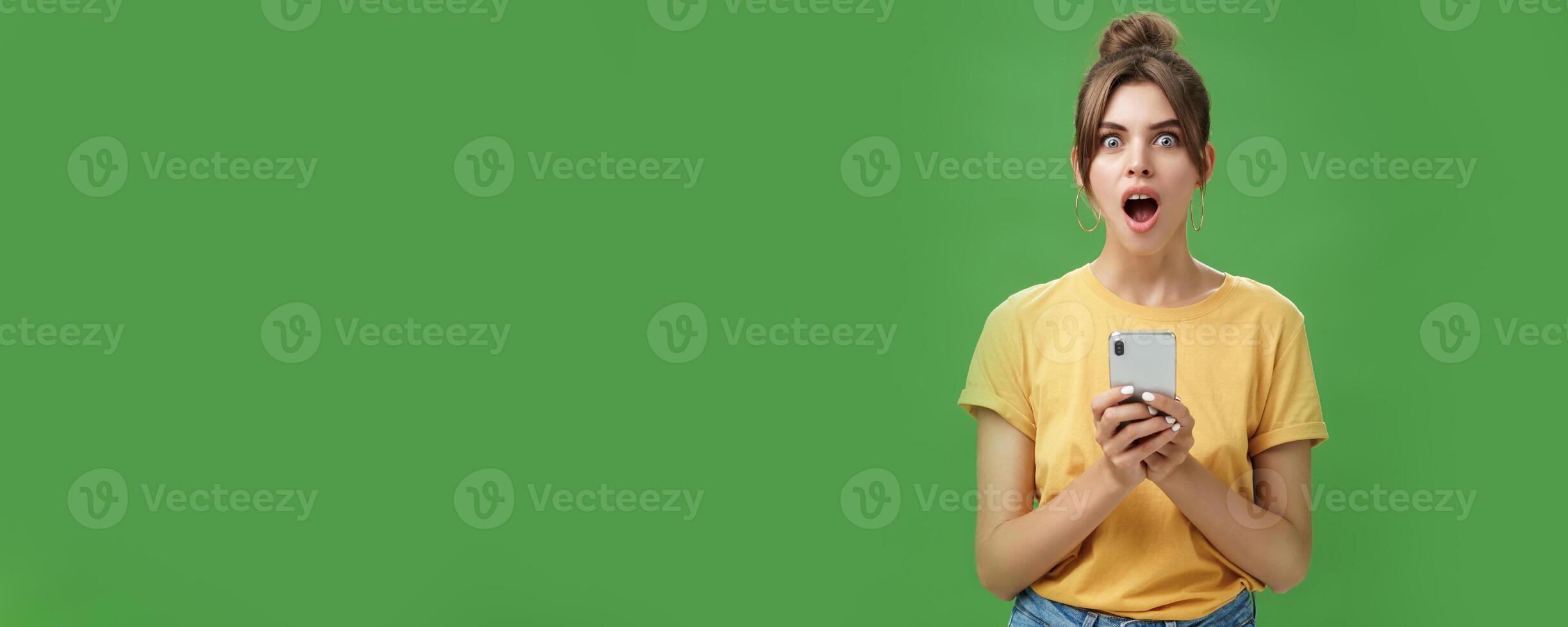 Portrait of shocked speechless and impressed beautiful white girl with combed hair in yellow t-shirt holding smartphone, dropping jaw from excitement reacting to cool app over green background photo