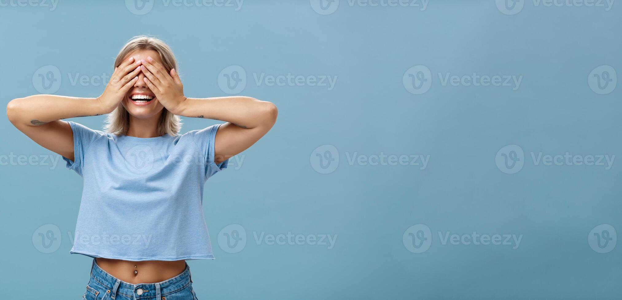 Hide if you can. Portrait of carefree and happy good-looking playful girl with blonde hair smiling broadly while counting ten and playing hide-and-seek with closed eyes over palms near blue wall photo
