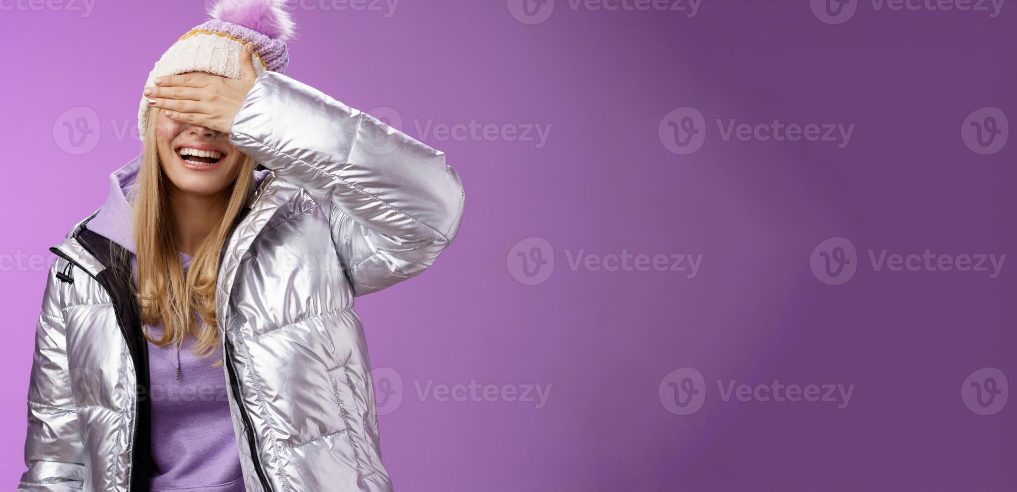 Happy relaxed smiling attractive young blond woman in stylish warm silver jacket hat hide eyes behind palm laughing happily waiting boyfriend prepared surprise gift amused, standing purple background photo