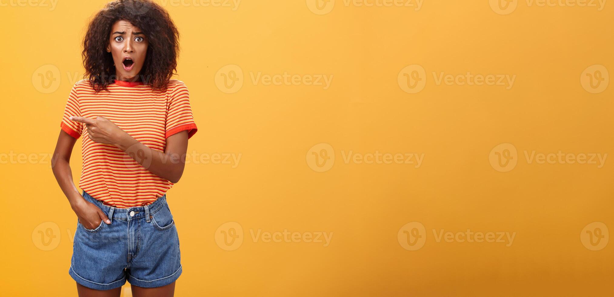 Studio shot of offended shocked stylish woman being robbed fasping and staring nervously begging stranger help out pointing left concerned and worried frowning, posing displeased over orange wall photo