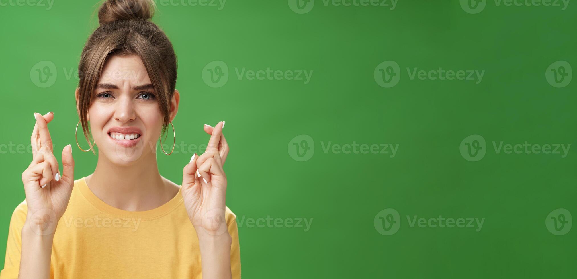 Close-up shot of nervous woman praying for having positive answer crossing fingers for good luck biting lower lip anxiously and frowning being worried while hoping to get job over green background photo