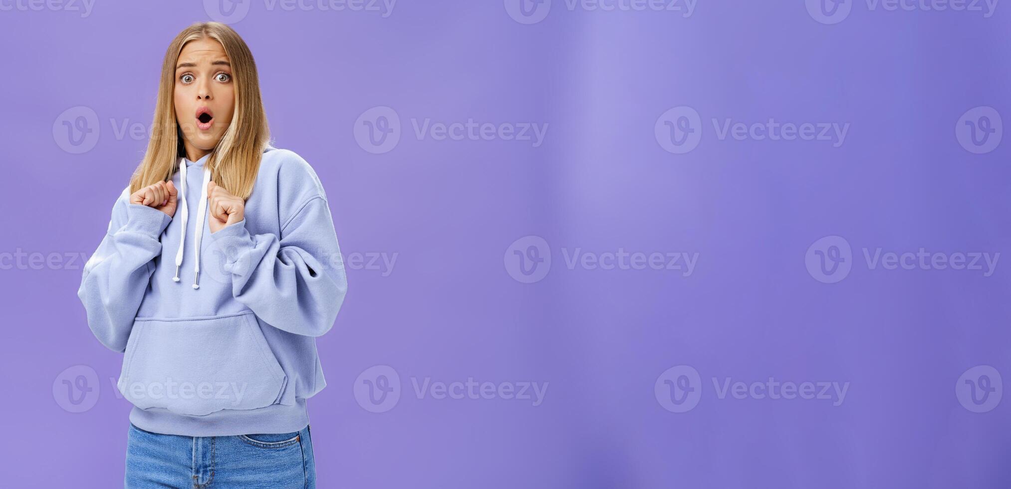 Woman scared of sudden loud noise in dark gasping looking shocked and concerned tilting backwards with frightened look pressing clenched fists to body from fear, wearing warm hoodie over purple wall photo