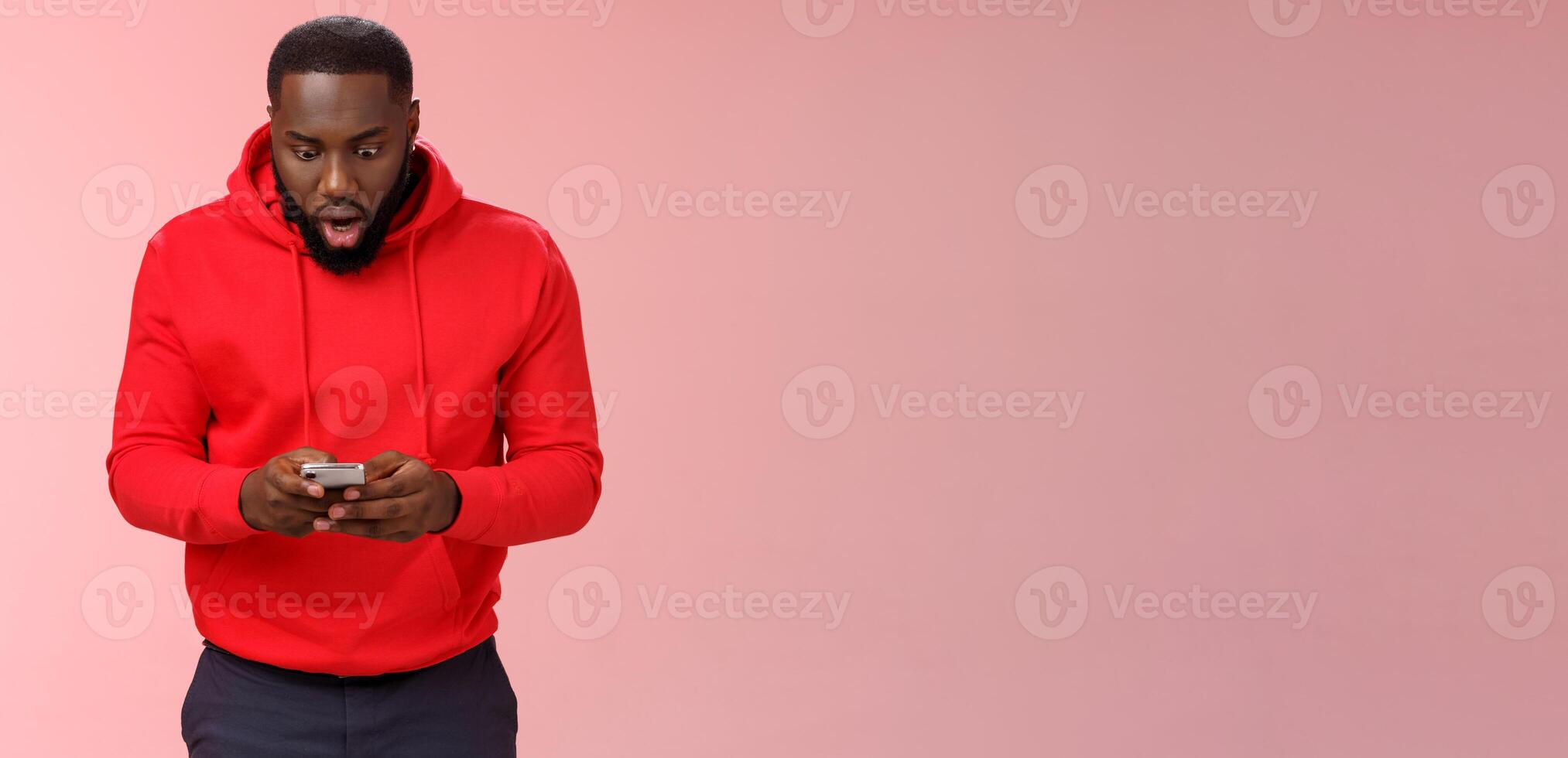 Amused focused impressed african-american bearded guy in red hoodie drop jaw widen eyes look amazed smartphone display winning hard game level answering exciting message, standing pink background photo