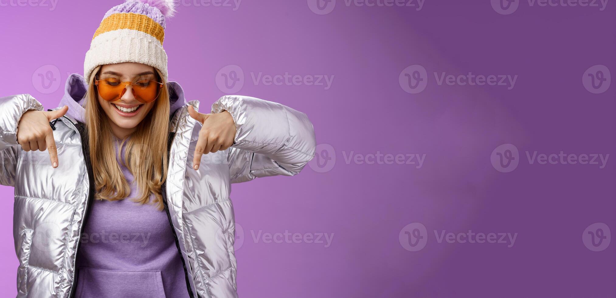 Joyful stylish blond woman in silver jacket stylish sunglasses jat enjoying perfect mountains view snowy vacation resort look pointing down smiling amused having fun feel happy, purple background photo