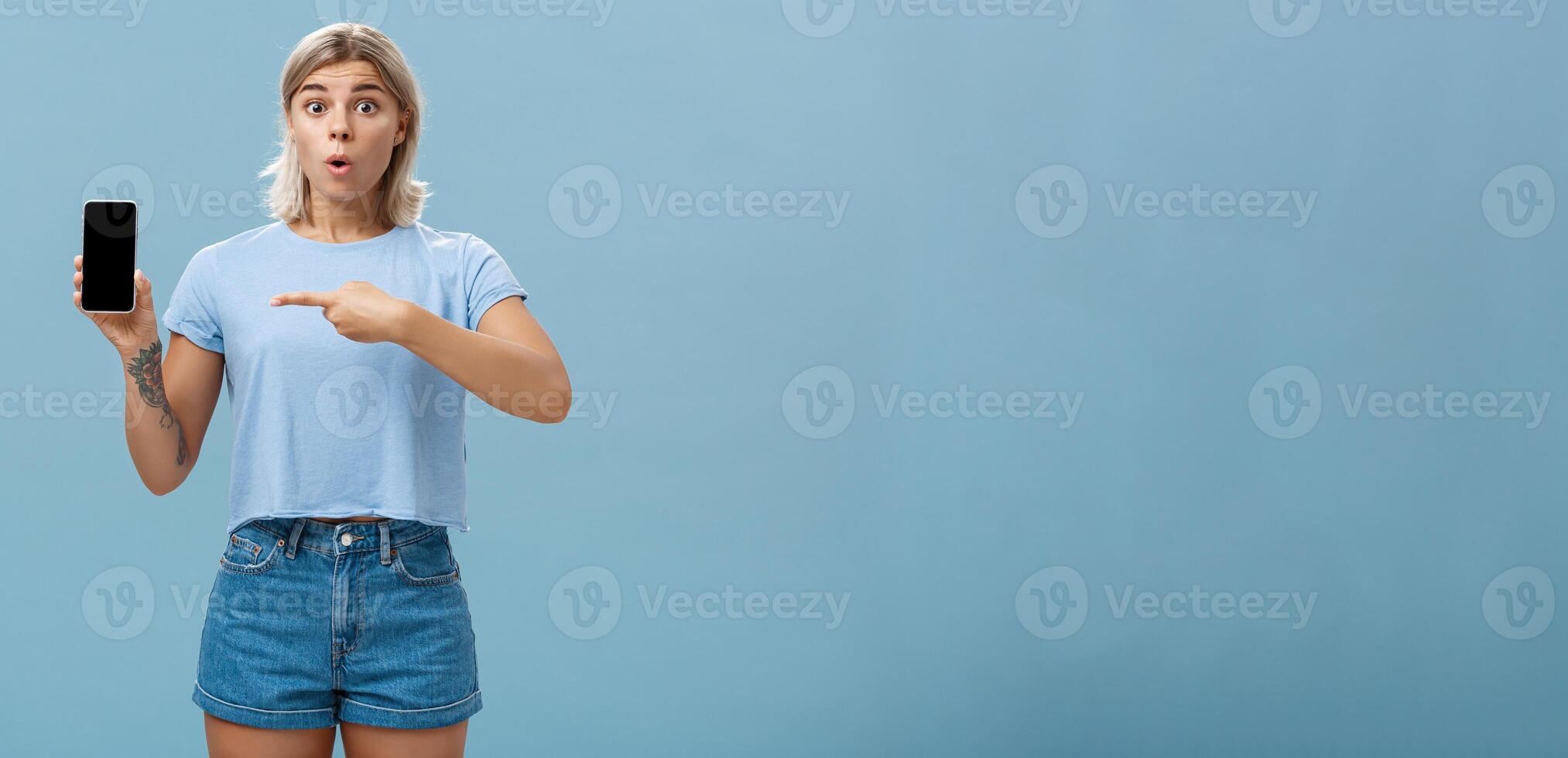 Wow look what found online. Portrait of impressed speechless good-looking young sportswoman in t-shirt and denim shorts saying wow folding lips showing smartphone and pointing at screen over blue wall photo