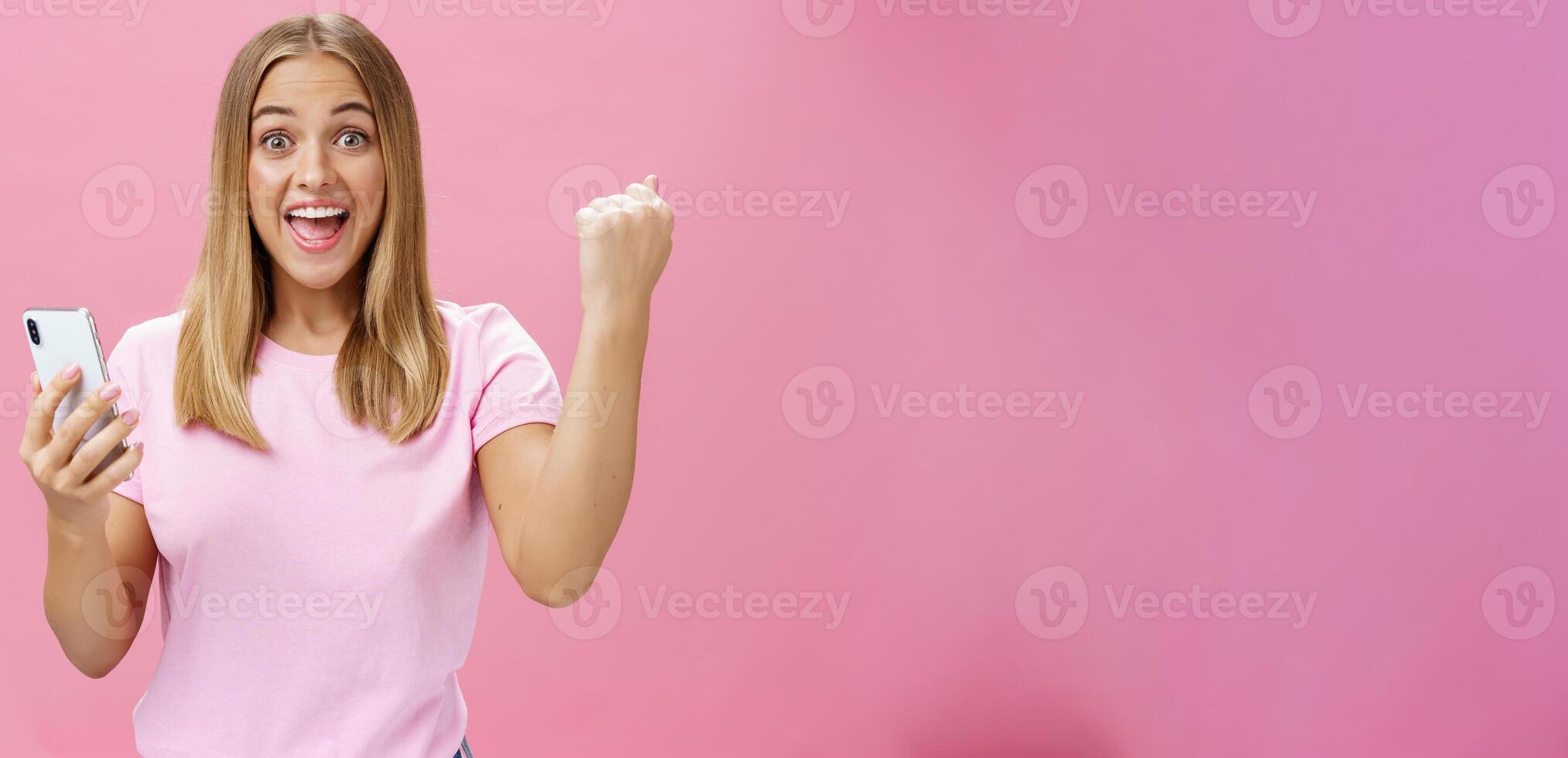 Girl beat own record in smartphone game raising clenched fist in cheer and triumph holding cellphone, smiling excited and happy at camera celebrating victory with joyful gesture over pink background photo
