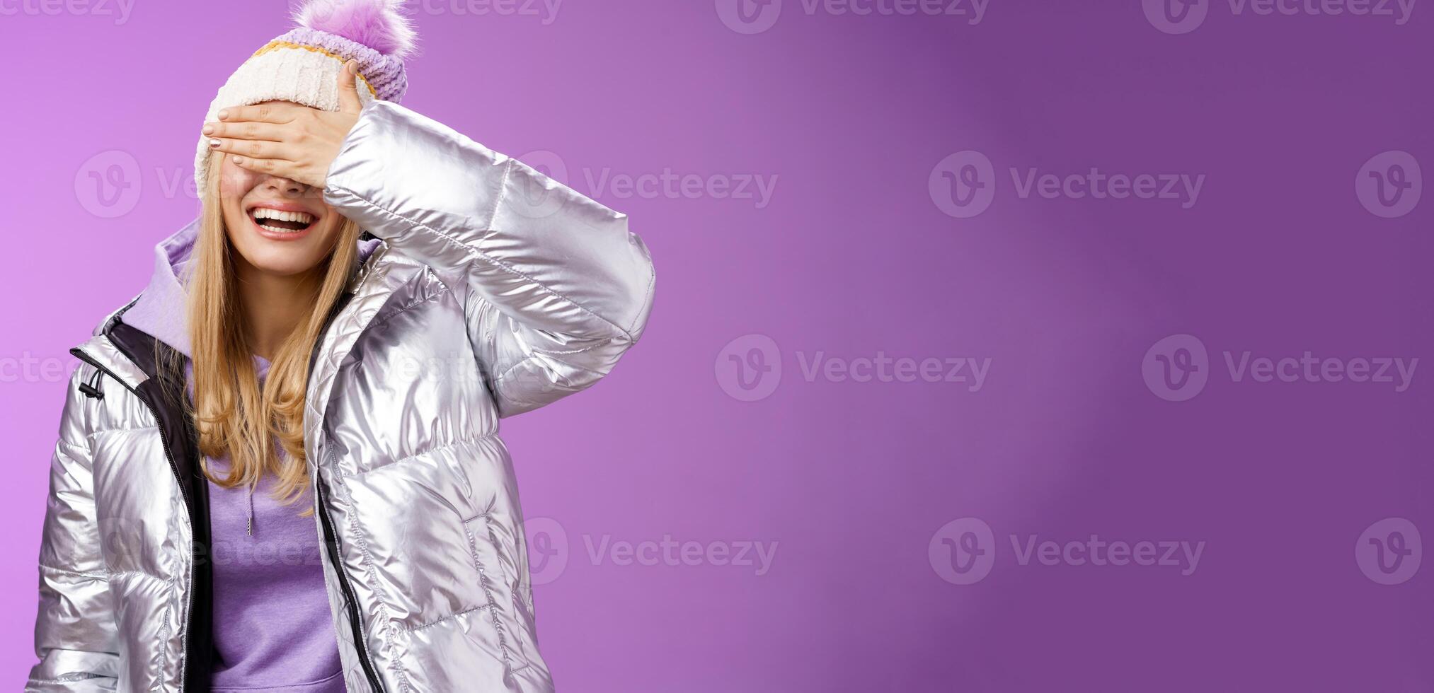 Happy relaxed smiling attractive young blond woman in stylish warm silver jacket hat hide eyes behind palm laughing happily waiting boyfriend prepared surprise gift amused, standing purple background photo