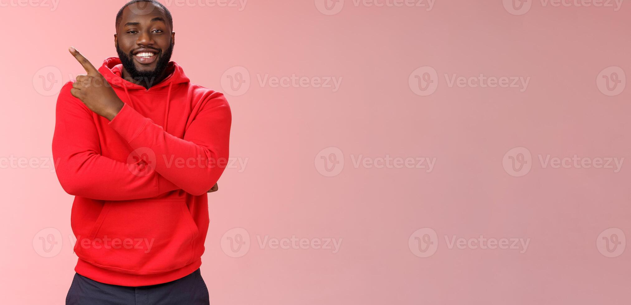 Cheeky outgoing smiling attractive african-american male model in red hoodie grinning energized pointing upper left corner indicating awesome product recommend promo, standing pink background photo