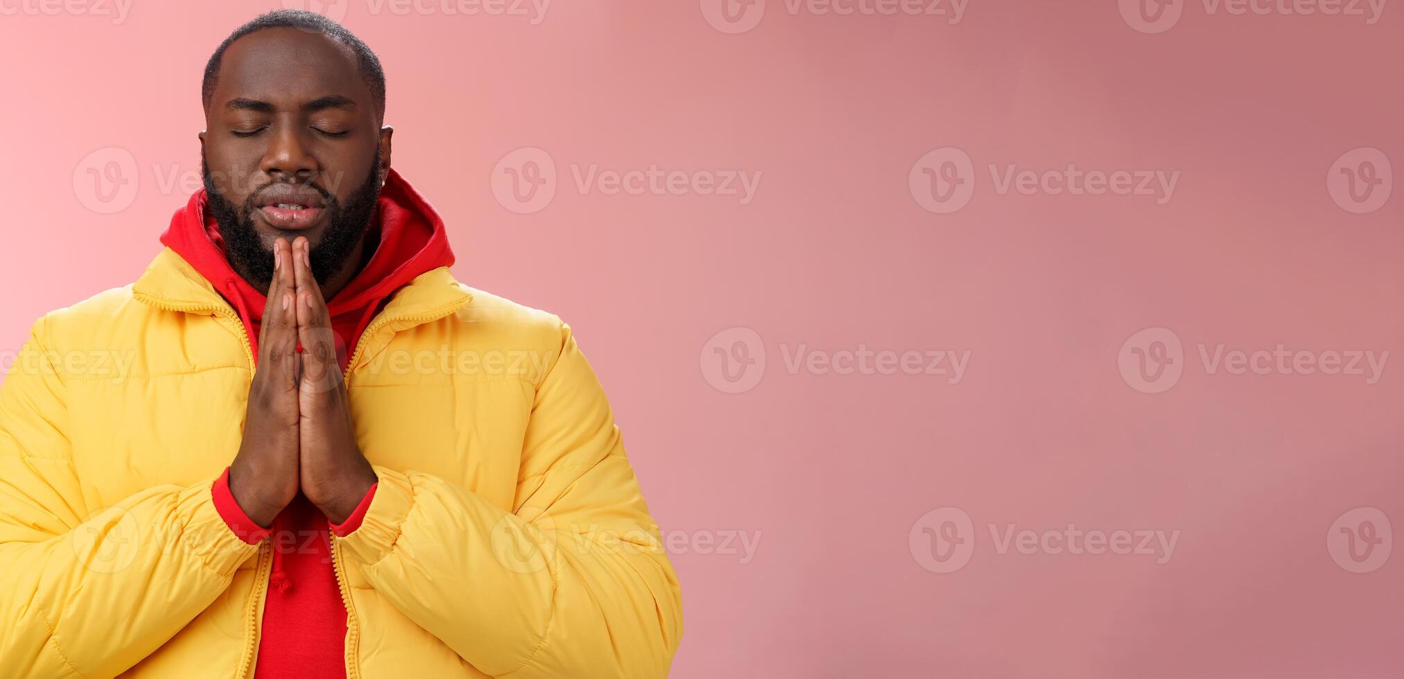 Serious-looking faithful young determined african-american praying guy in yellow coat press palms together pray close eyes whisper supplication asking god help make wish, standing pink background photo
